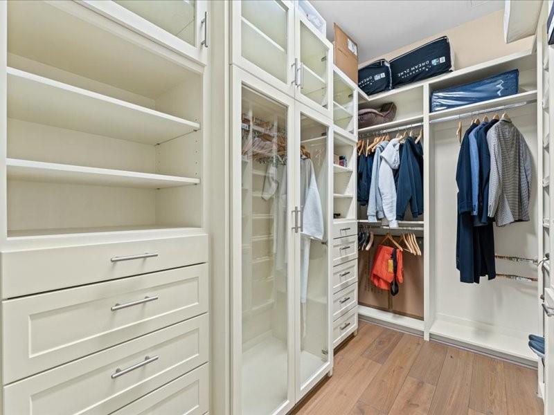 Walk-in closet with white built-ins, glass-front cabinets, hanging clothes, and wood flooring.