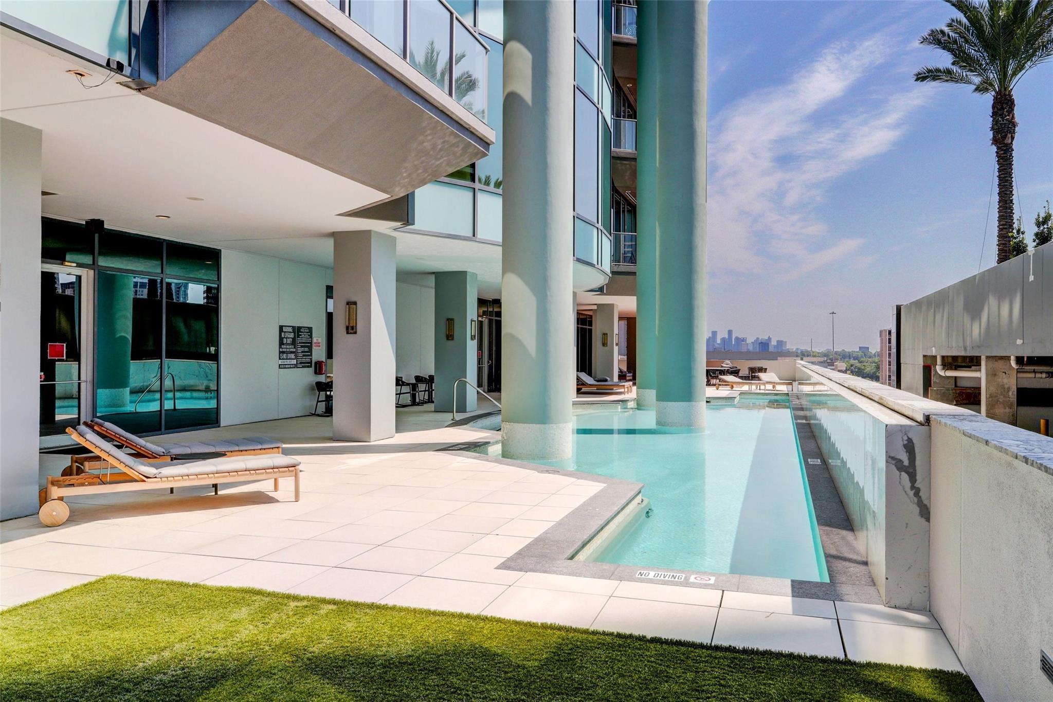 Rooftop pool with turquoise water, lounge chairs, tall columns, and a distant Houston skyline.