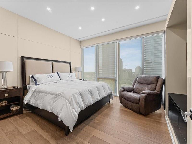 Bedroom with white bedding, floor-to-ceiling windows, wood floors, and a brown recliner.