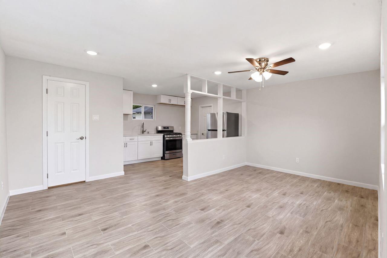 Vacant living room with wood-look tile flooring, ceiling fan, and open kitchen with stainless steel appliances.