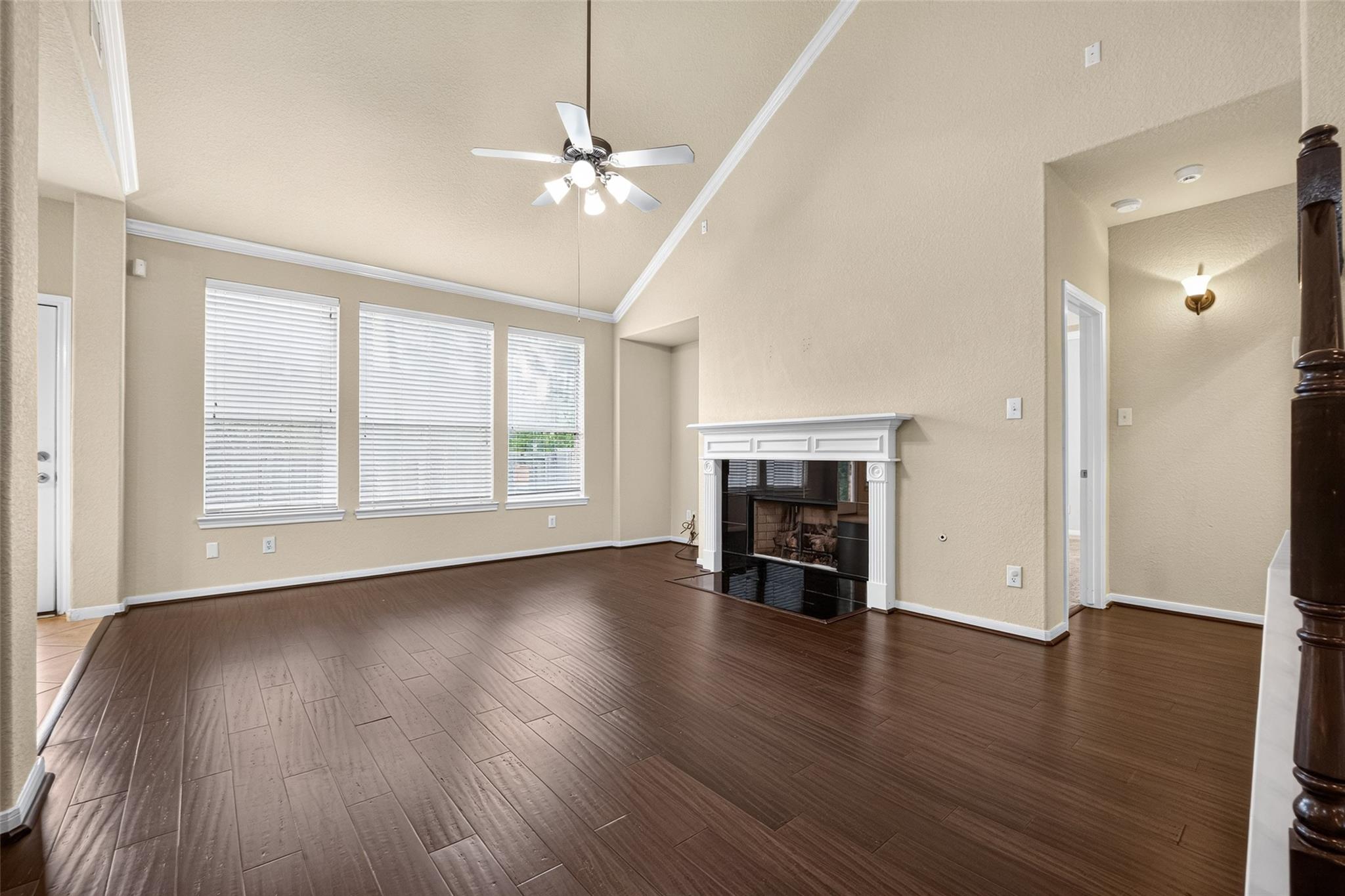 Empty living room with dark flooring, a white fireplace, three blinded windows, and a ceiling fan.