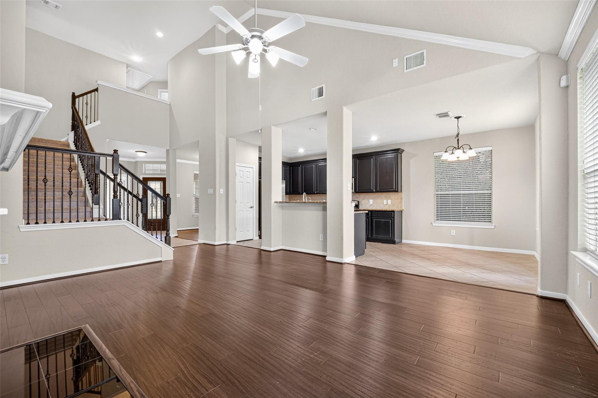 Open-plan living and dining area with dark wood floors, a tall ceiling, wrought-iron staircase railing, and tiled kitchen.