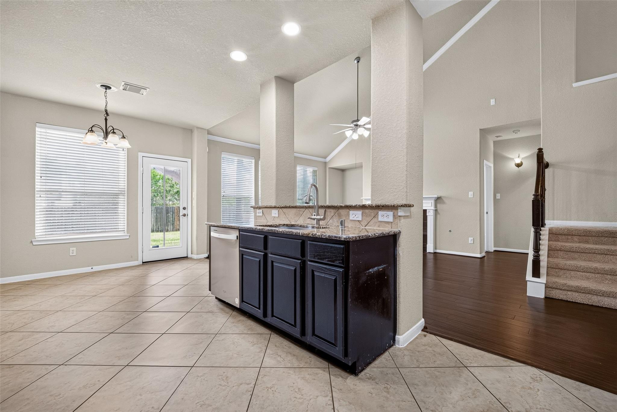 Open-plan kitchen with a dark island, granite counter, stainless dishwasher, and tile flooring beside a wood-floored hall.