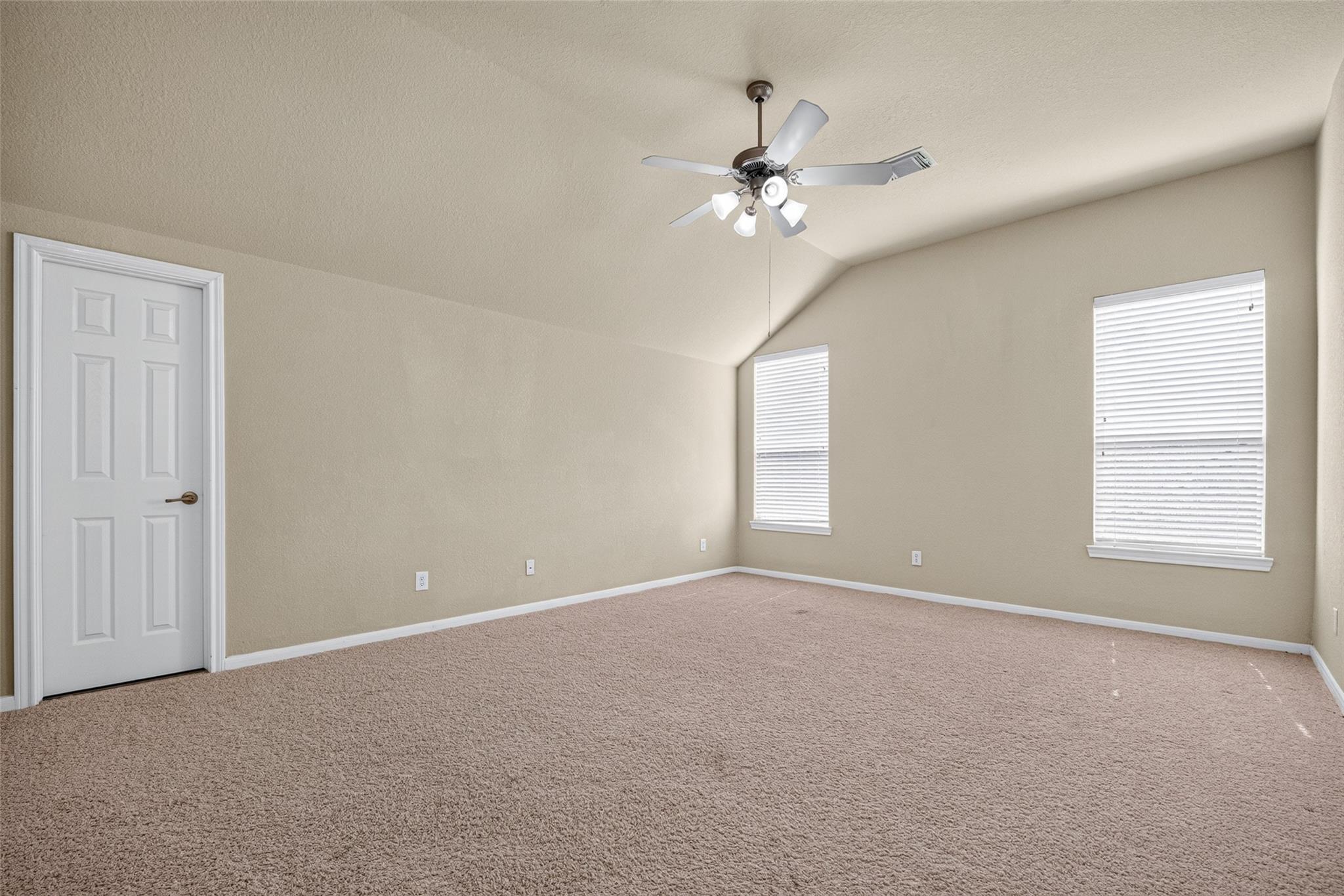 Empty carpeted room with a vaulted ceiling, ceiling fan, and two windows with blinds.