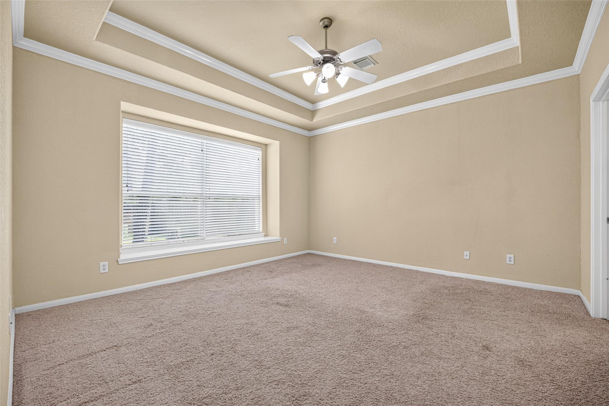 Empty carpeted room with a tray ceiling, ceiling fan, and large window with blinds.