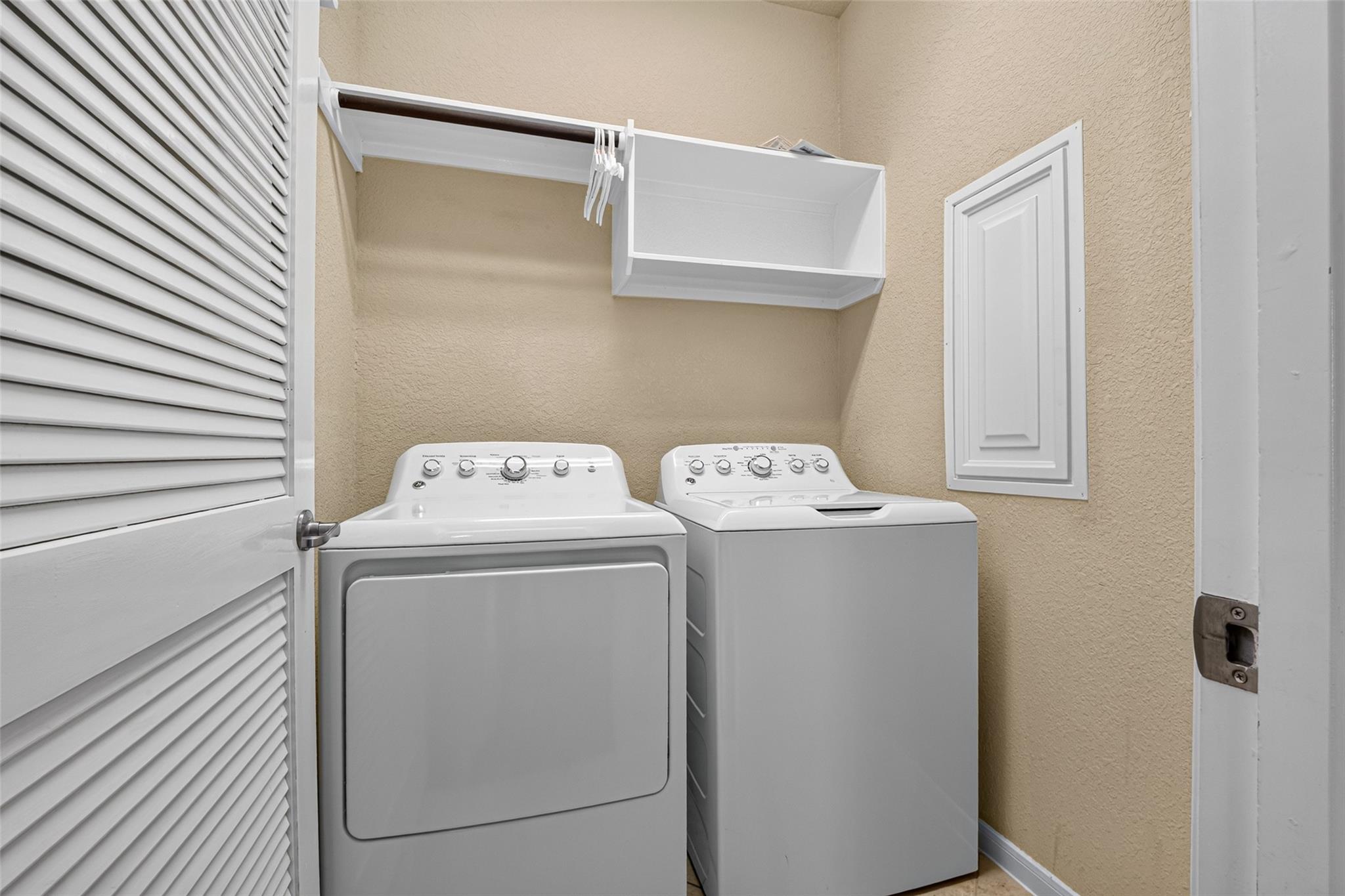 Laundry room with a dryer, top-loading washer, and overhead storage shelf.