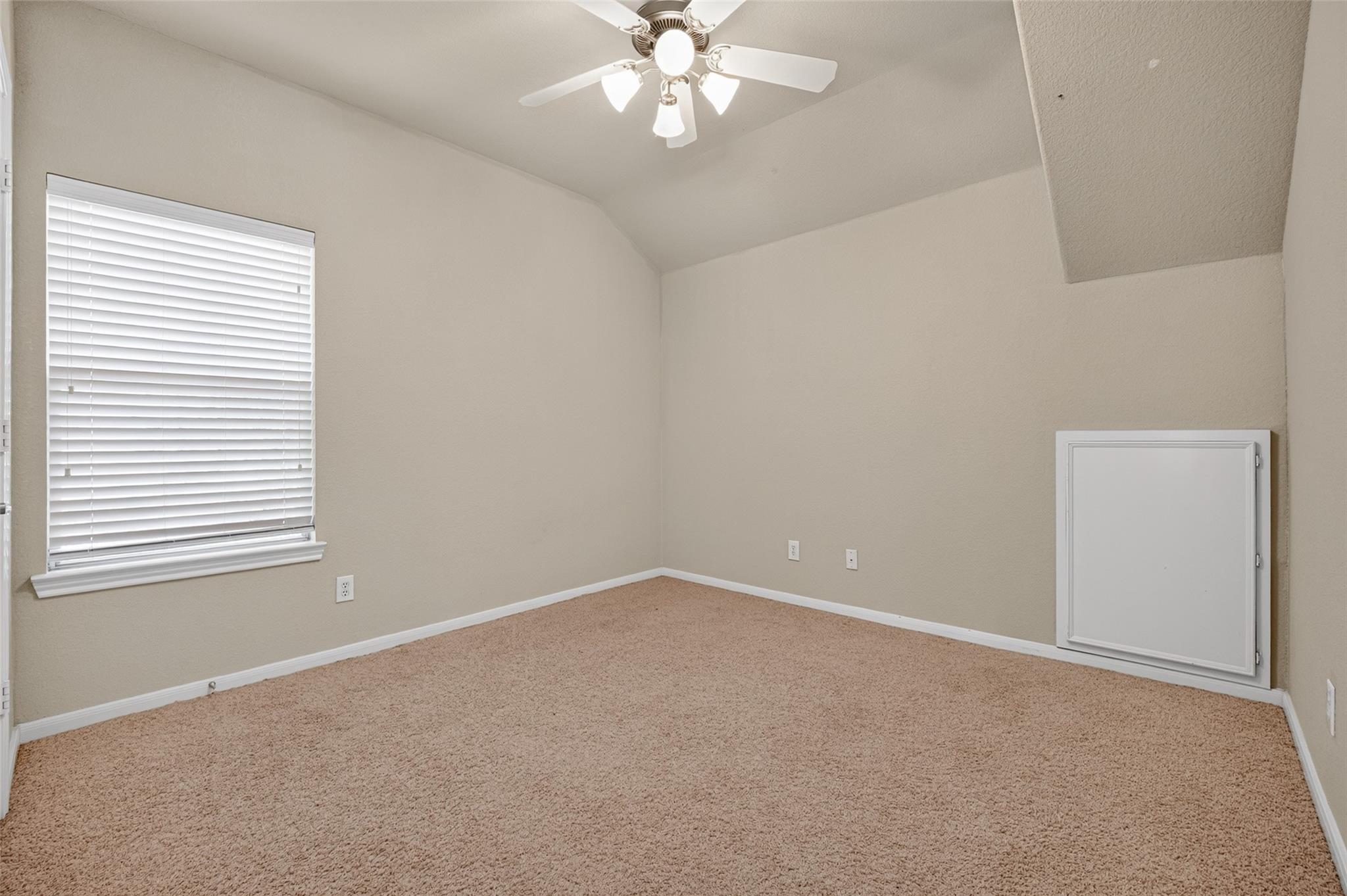 Empty carpeted room with a window, ceiling fan, and small wall access door.