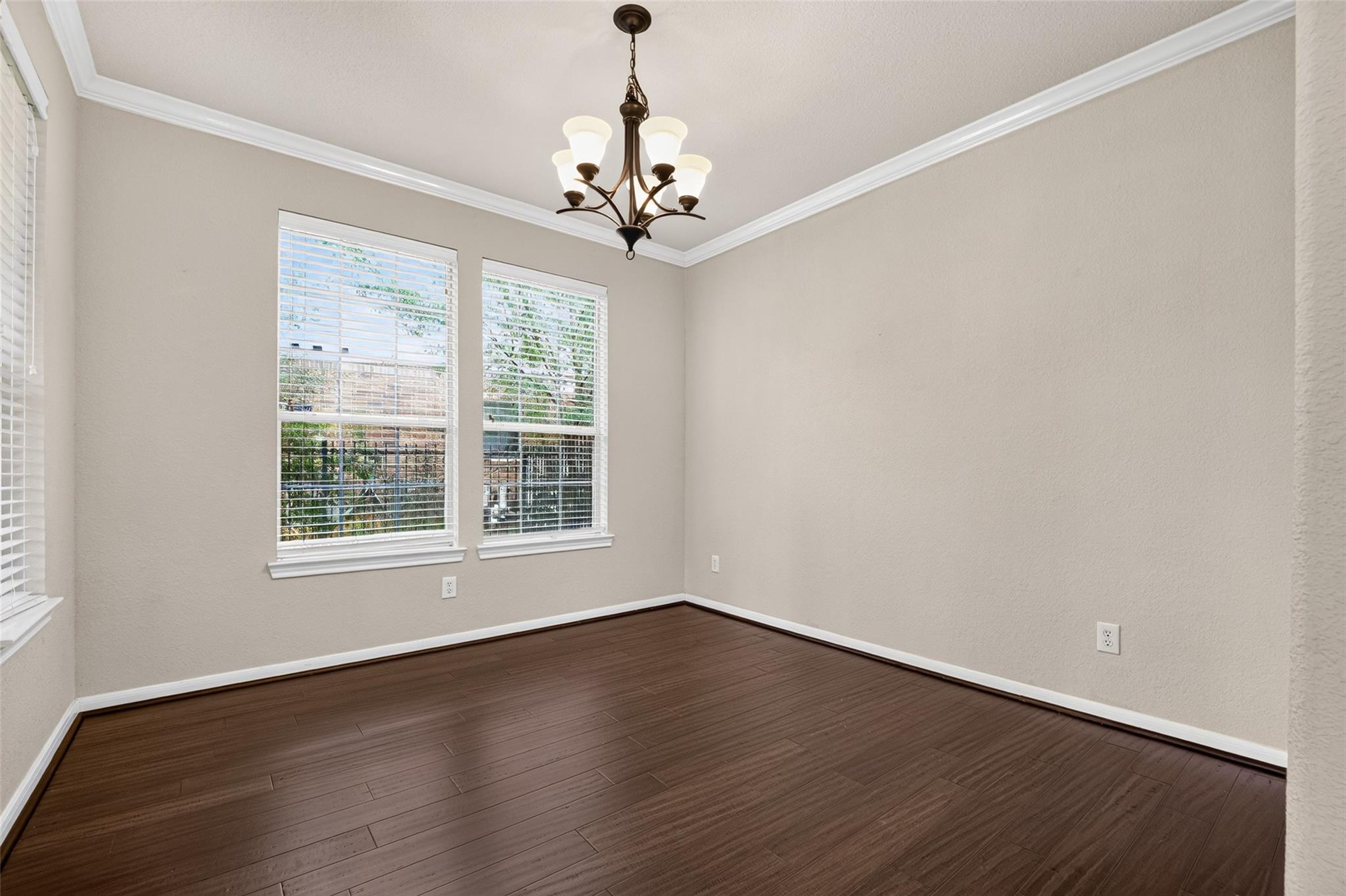 Empty dining room with dark wood floors, two blinded windows, and a hanging chandelier.