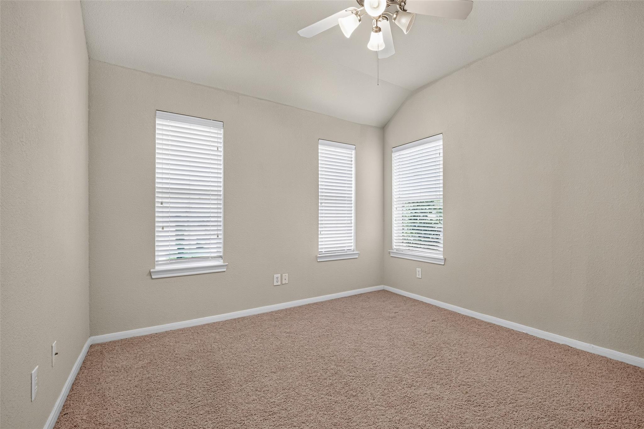 Empty bedroom with beige carpet, vaulted ceiling, three windows, and ceiling fan