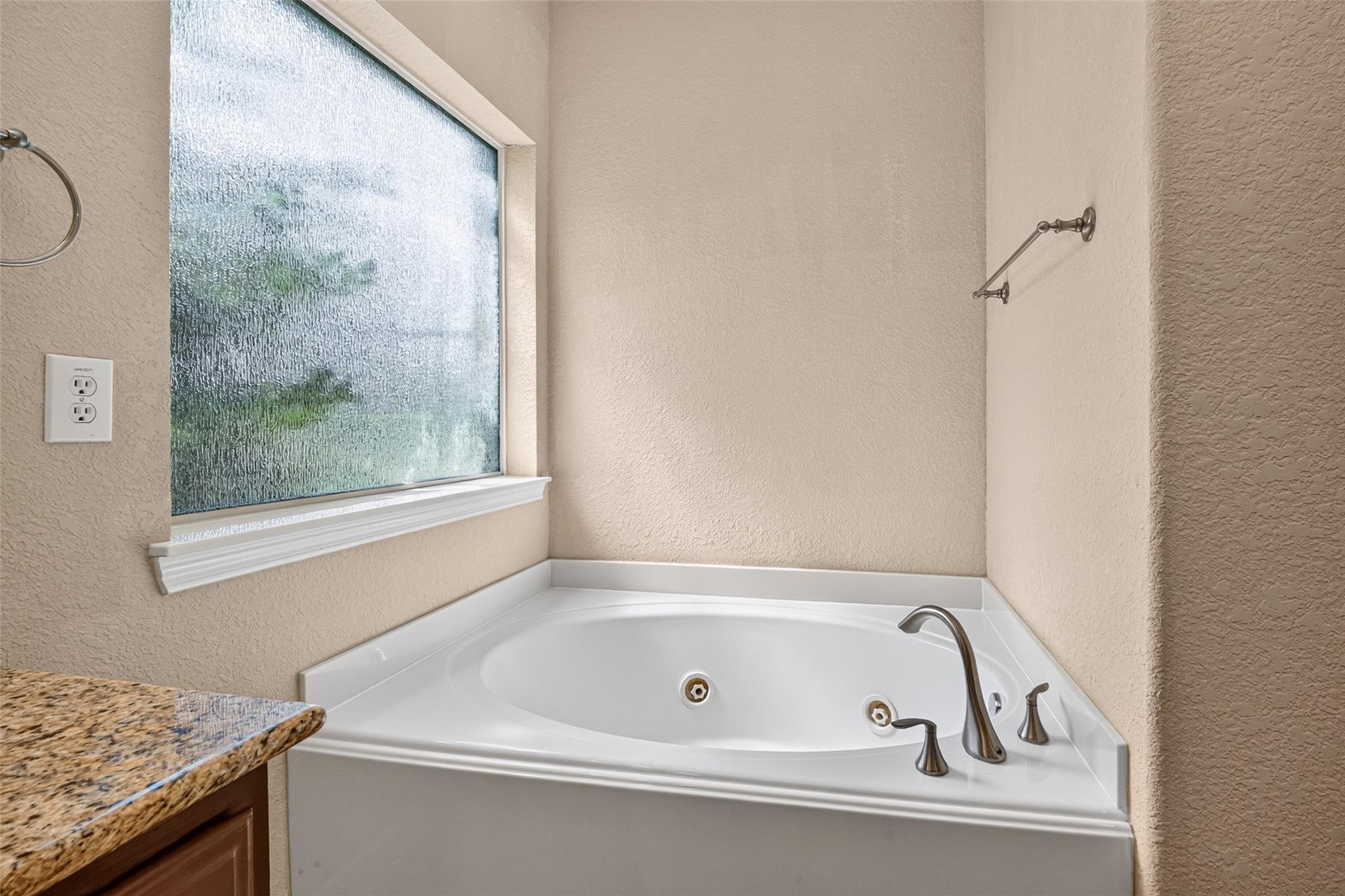 Corner jetted bathtub beneath a frosted window, with chrome fixtures and a granite countertop edge.