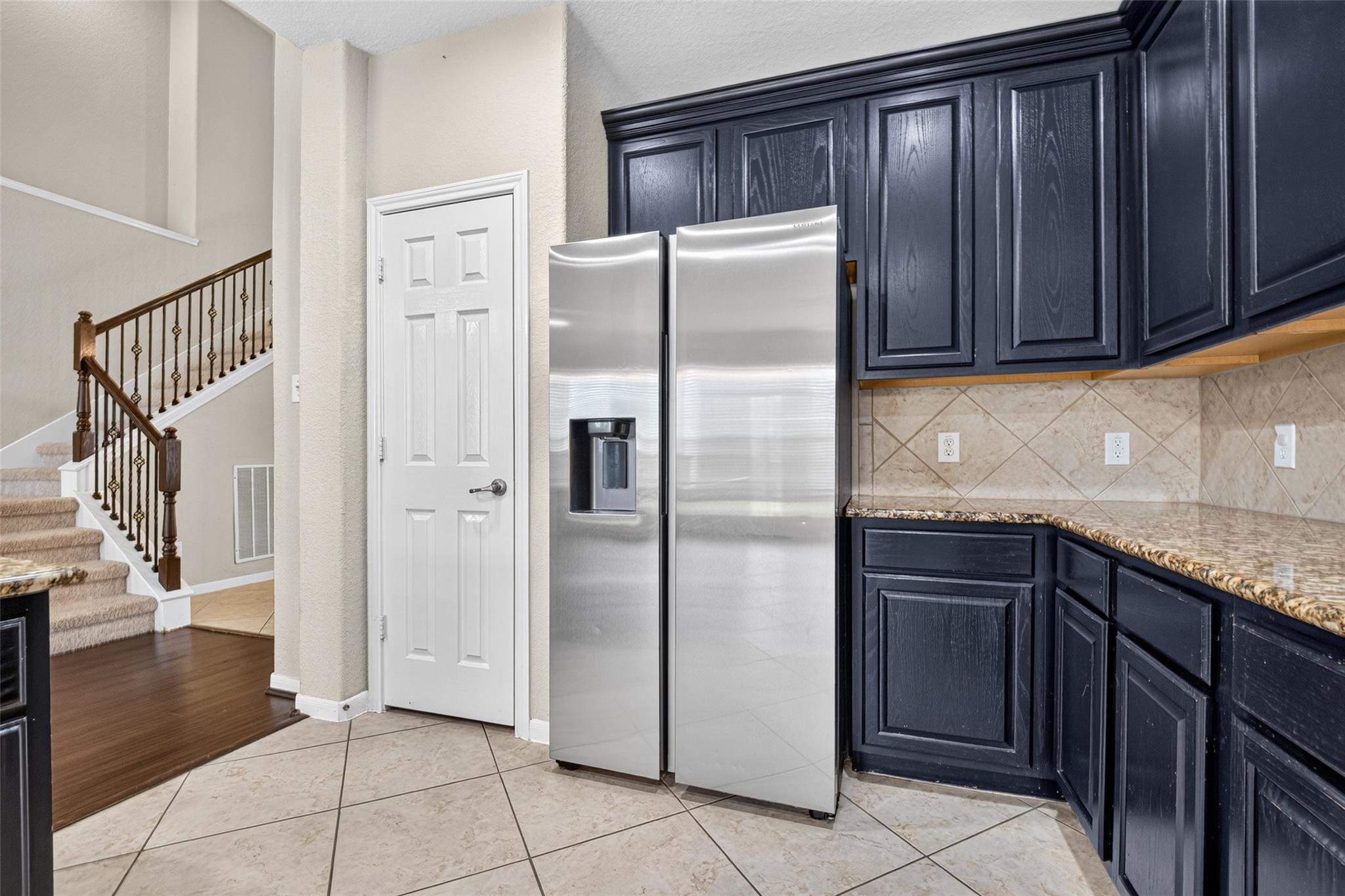 Kitchen with dark blue cabinets, stainless steel refrigerator, granite counters, and a staircase alongside.