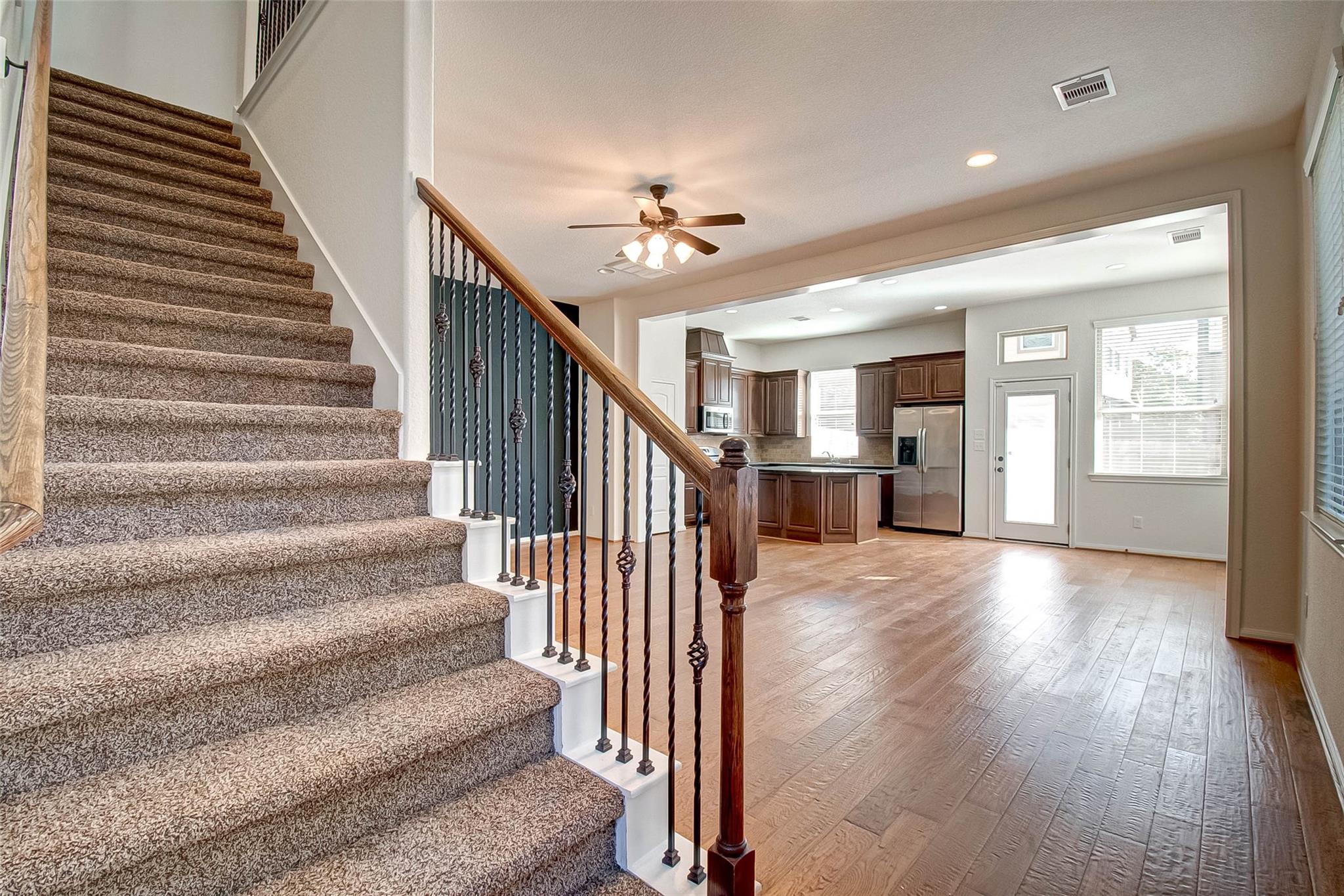 Unfurnished entry with a carpeted staircase, iron balusters, and an open kitchen with dark cabinets.