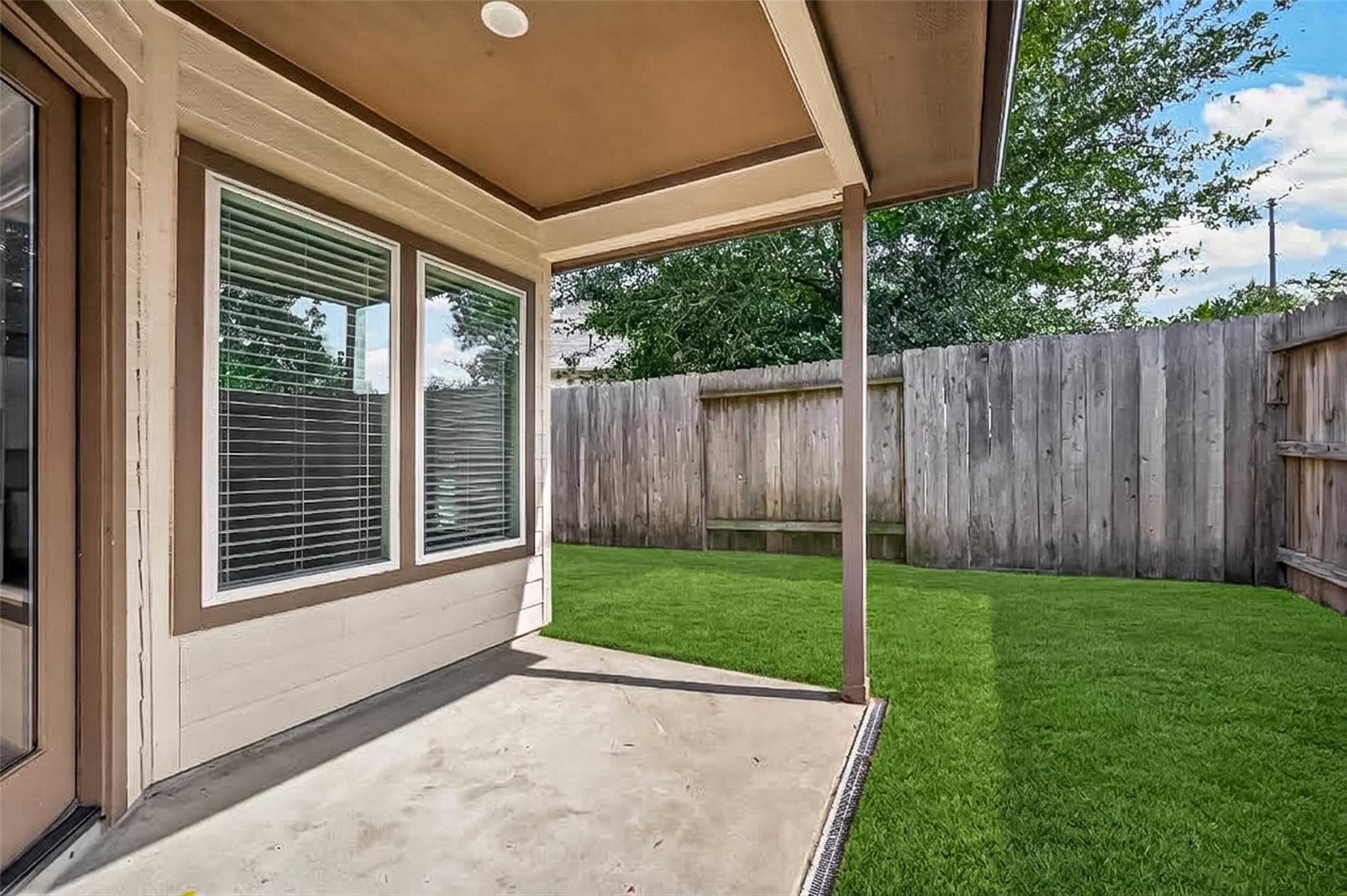 Covered concrete patio beside a fenced backyard with green lawn and mature trees.