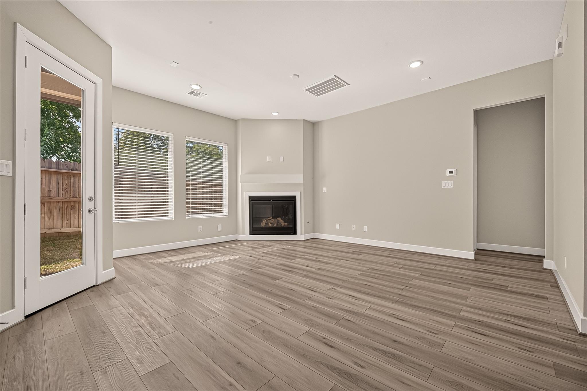 Empty living room with wood-look flooring, a corner fireplace, and windows beside a glass patio door.