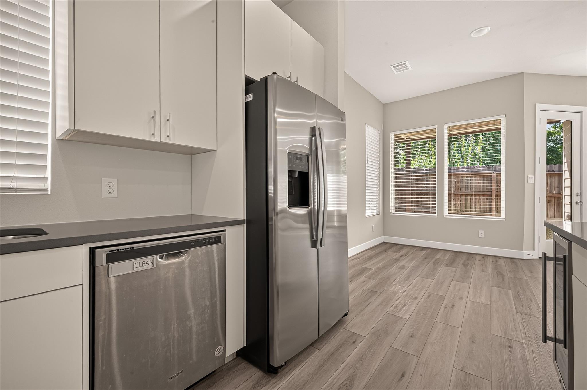 Kitchen with stainless steel appliances, white cabinets, and wood-look flooring beside windows overlooking a fenced yard.