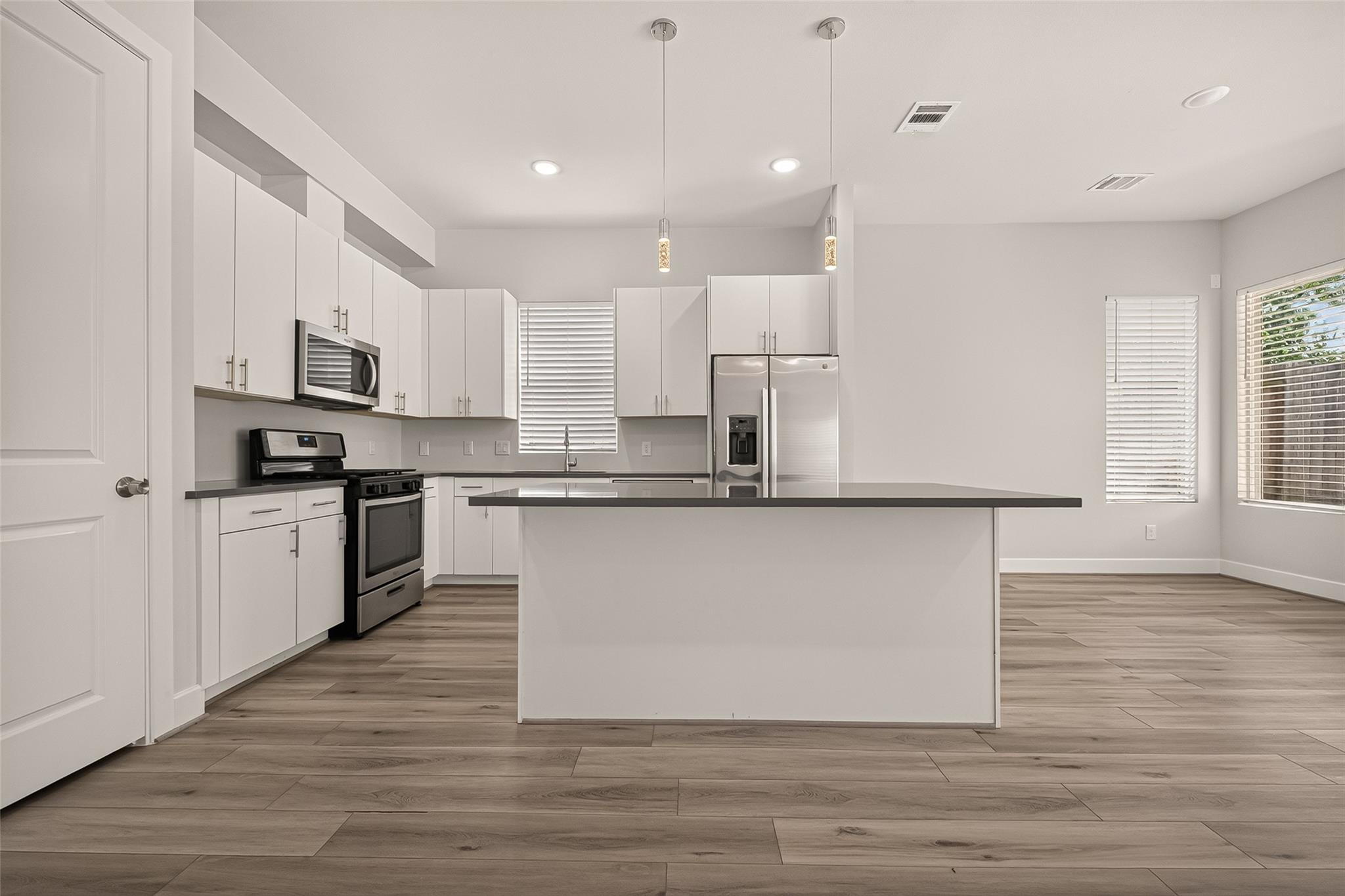 Vacant kitchen with a large island, white cabinetry, stainless steel appliances, and wood-look tile flooring.