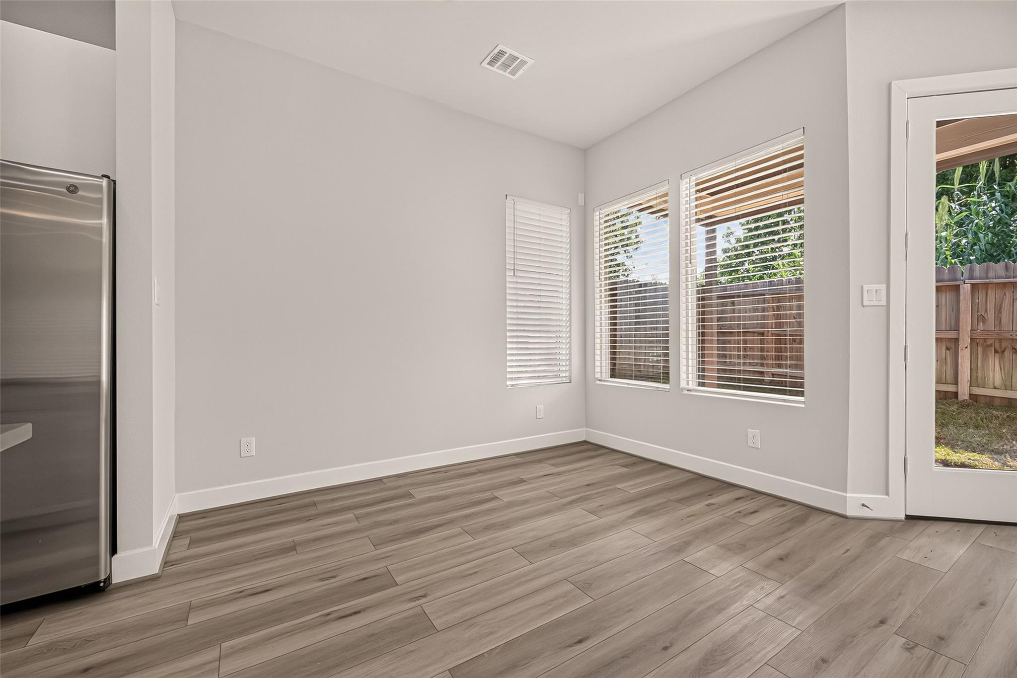 Empty kitchen area with wood-look floors, a stainless refrigerator, and windows overlooking a fenced yard.