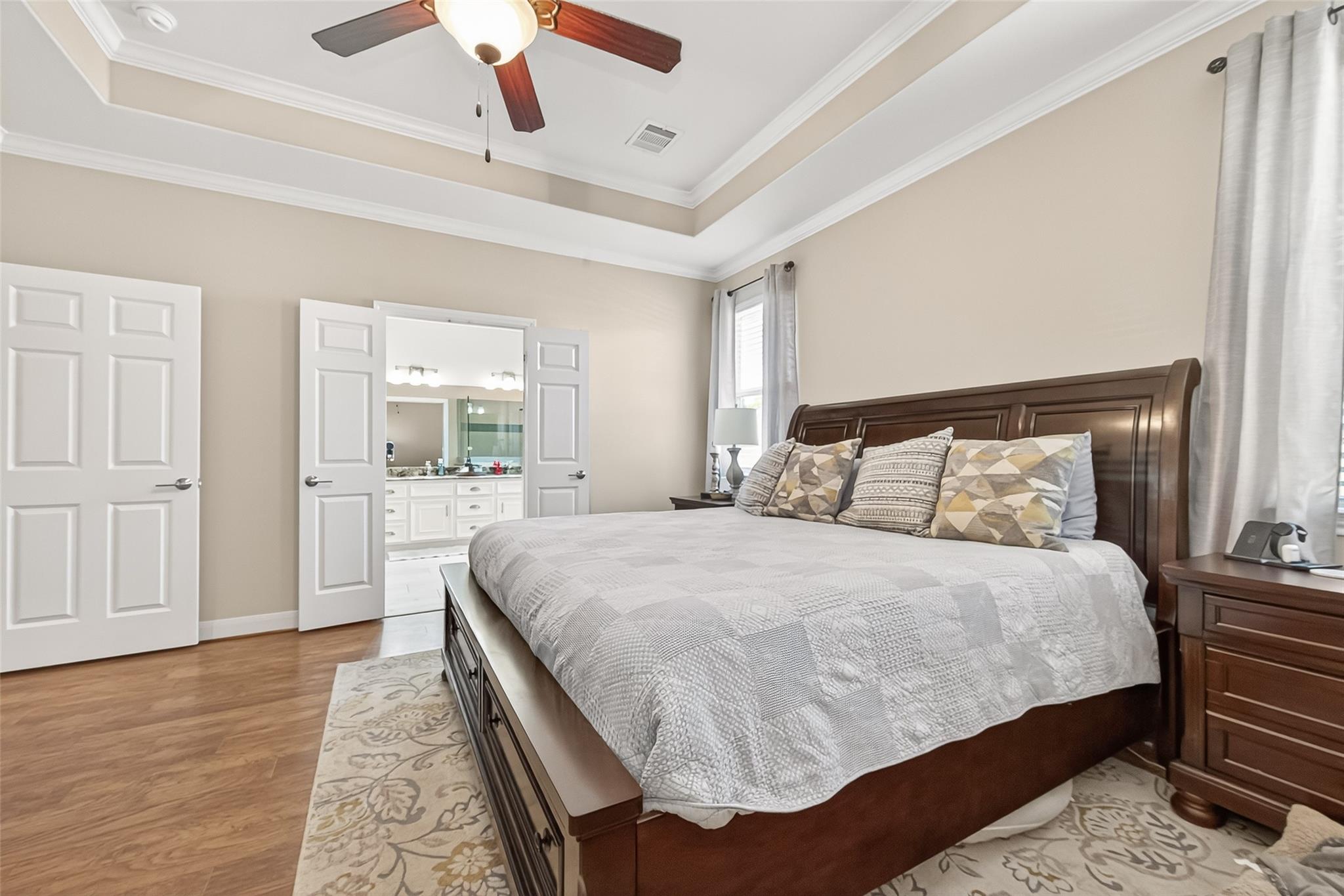 Primary bedroom with a dark wood bed, tray ceiling, and open doors to the adjoining bathroom.