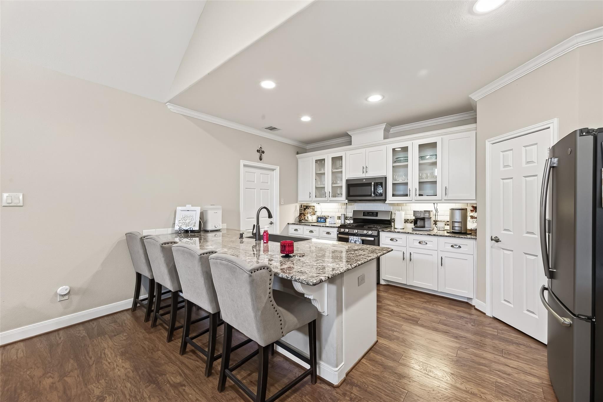 Kitchen with white cabinetry, stainless steel appliances, granite island, and four upholstered bar stools.