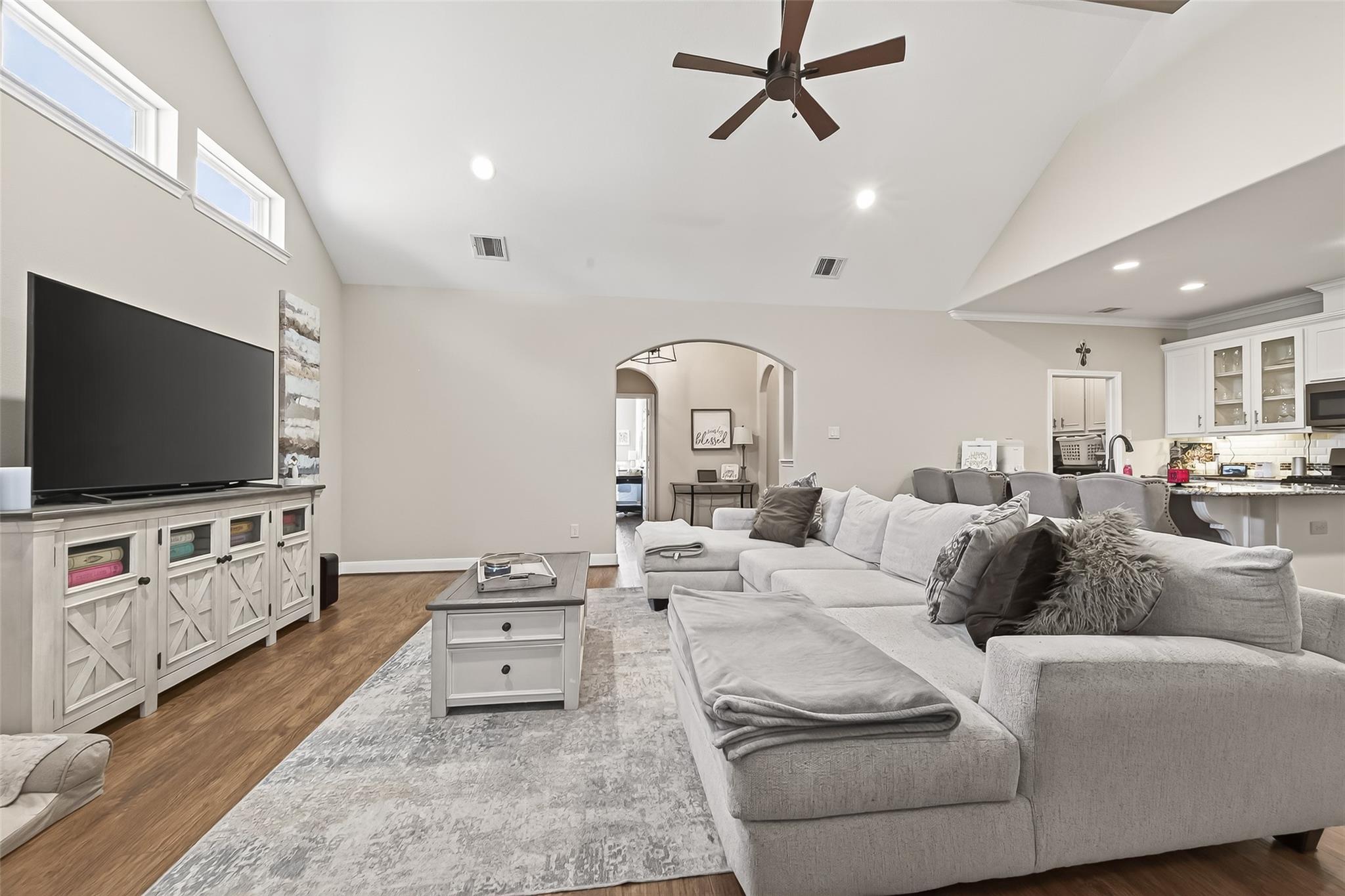 Living room with a vaulted ceiling, gray sectional, high windows, and an open kitchen.