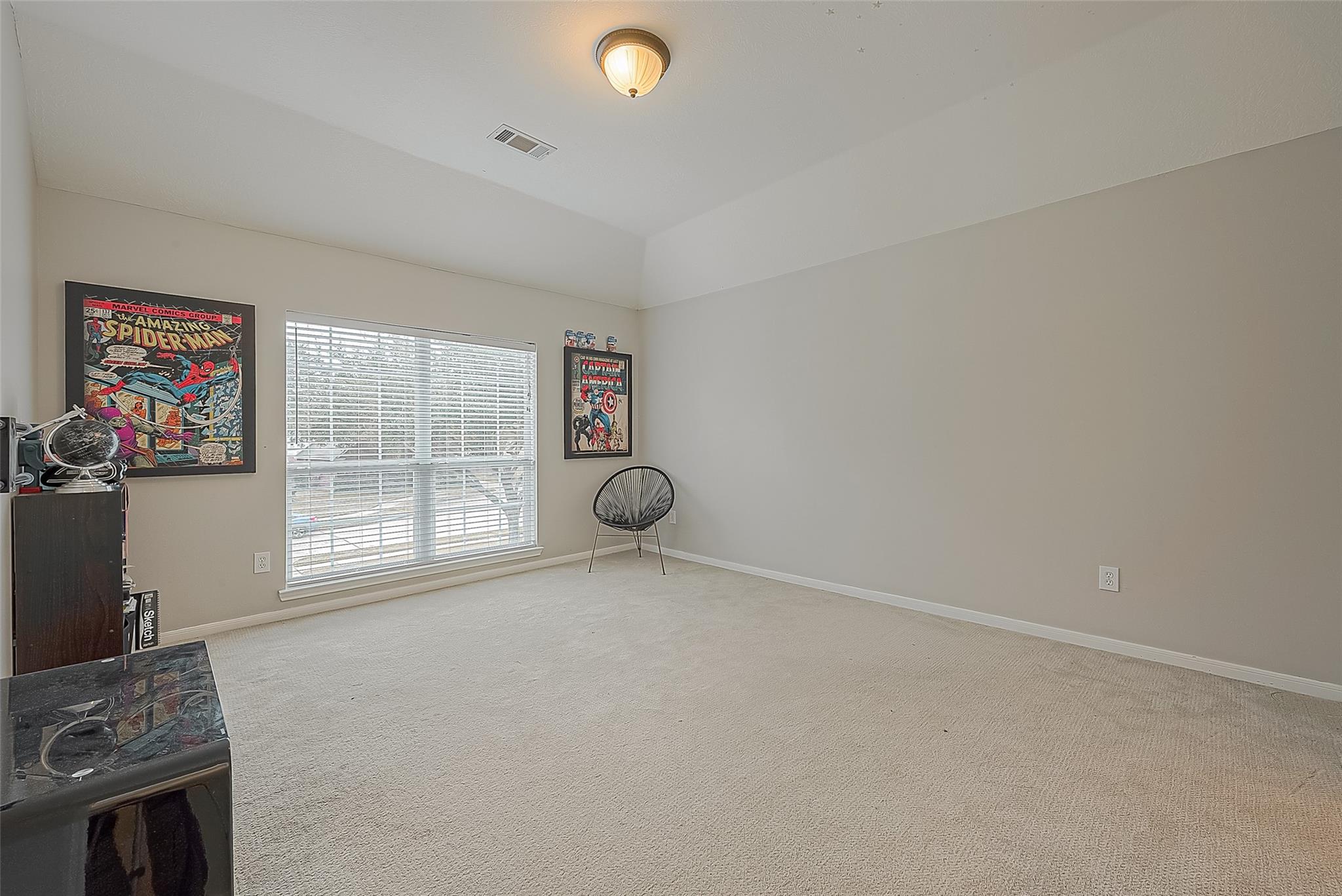 Carpeted room with a high ceiling, window blinds, framed comic art, and a black wire chair.