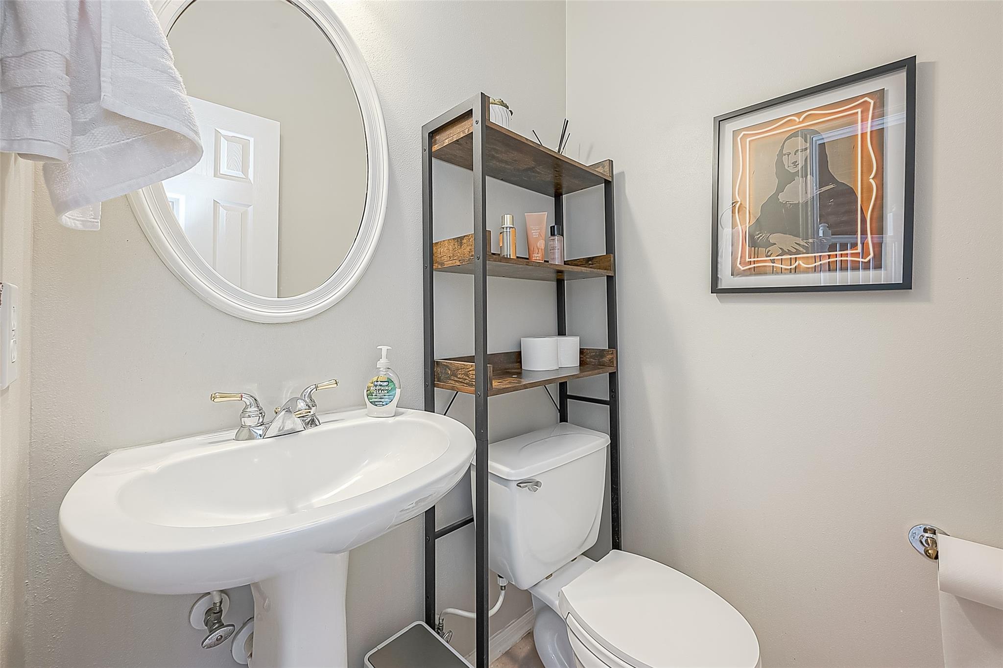 Bathroom with a pedestal sink, round mirror, and open shelving beside the toilet.