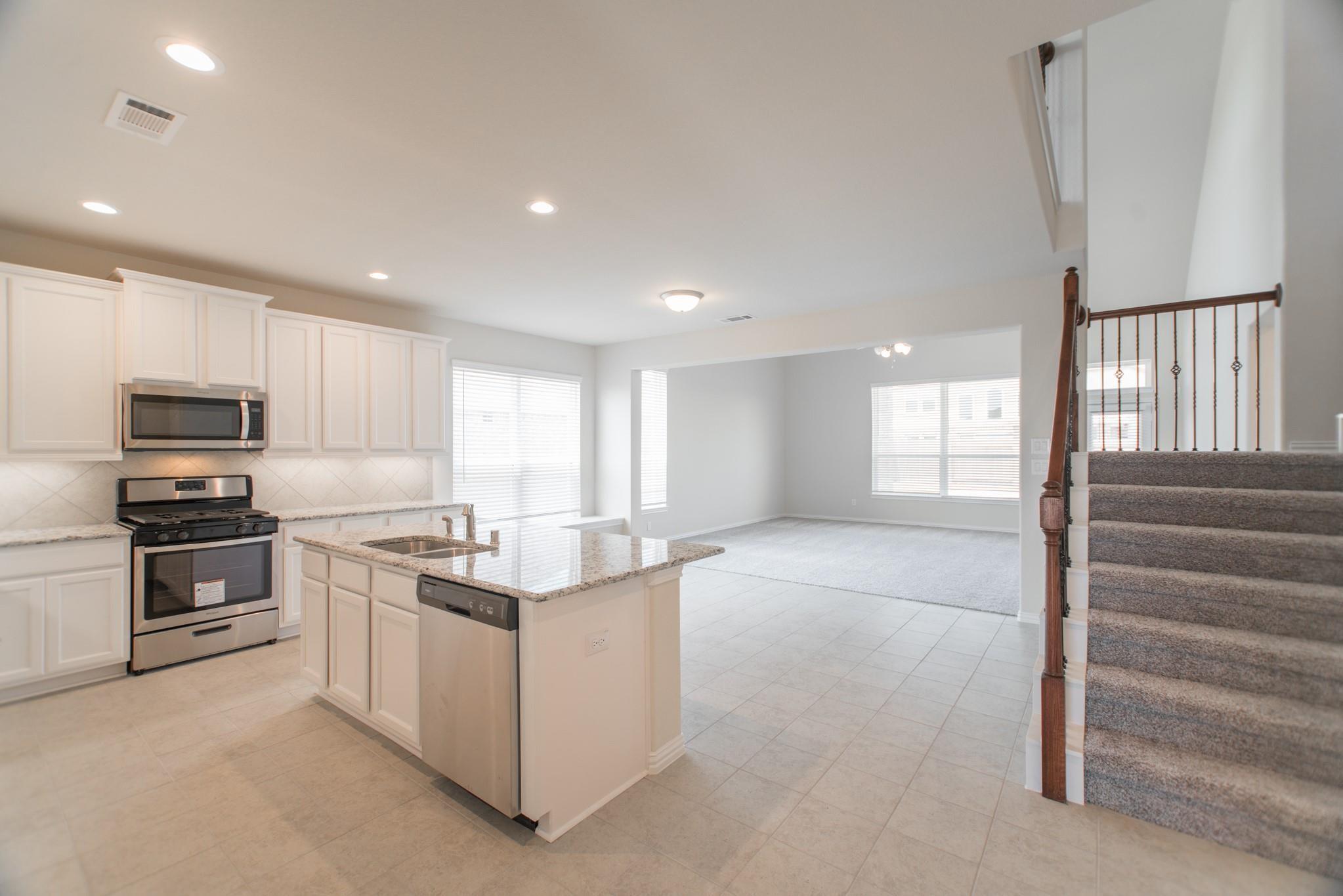 Open-plan kitchen with white cabinets, granite island, stainless appliances, and staircase beside the living area.