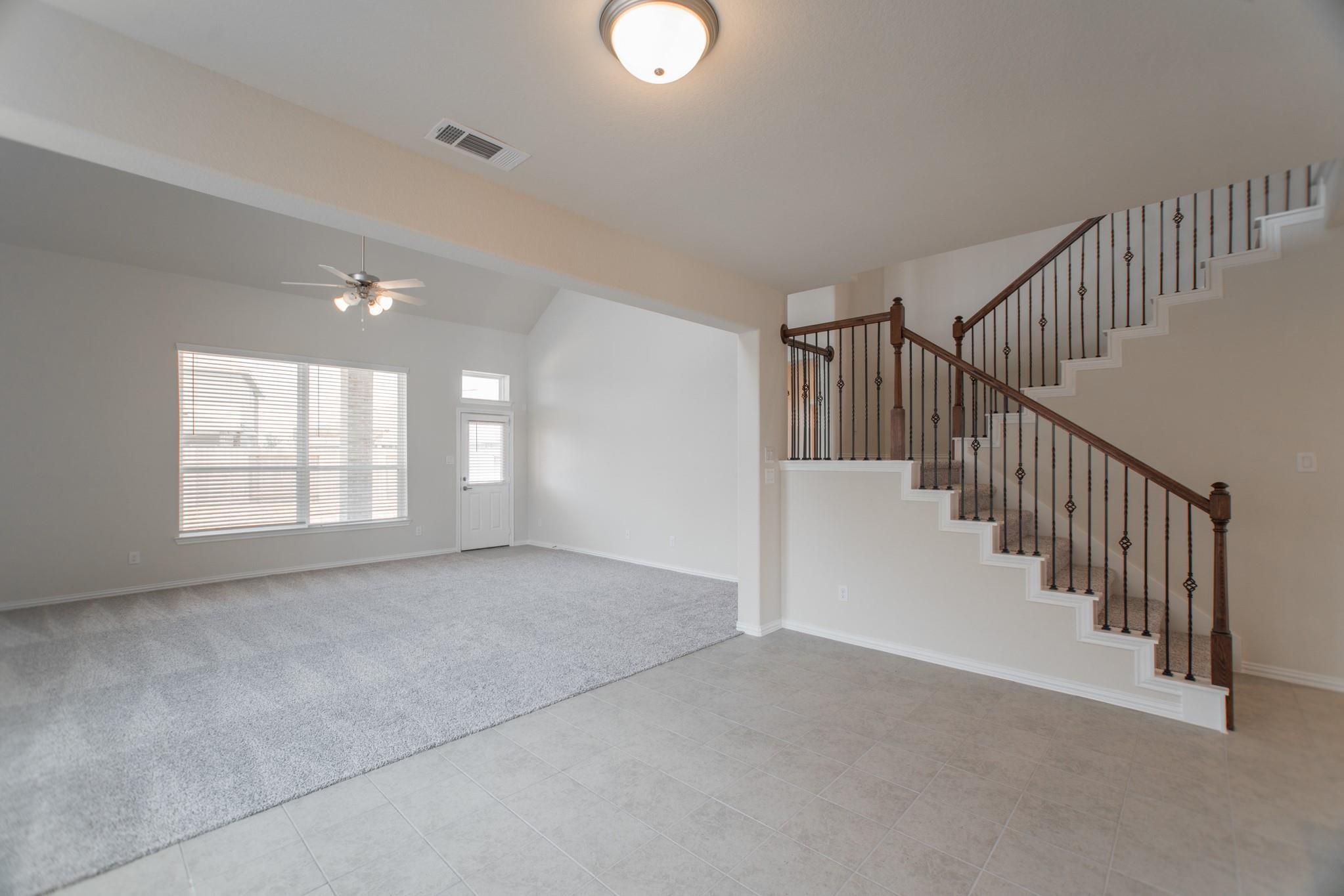 Open living area with gray carpet, tiled entry, and a staircase with dark wood rails and black balusters.