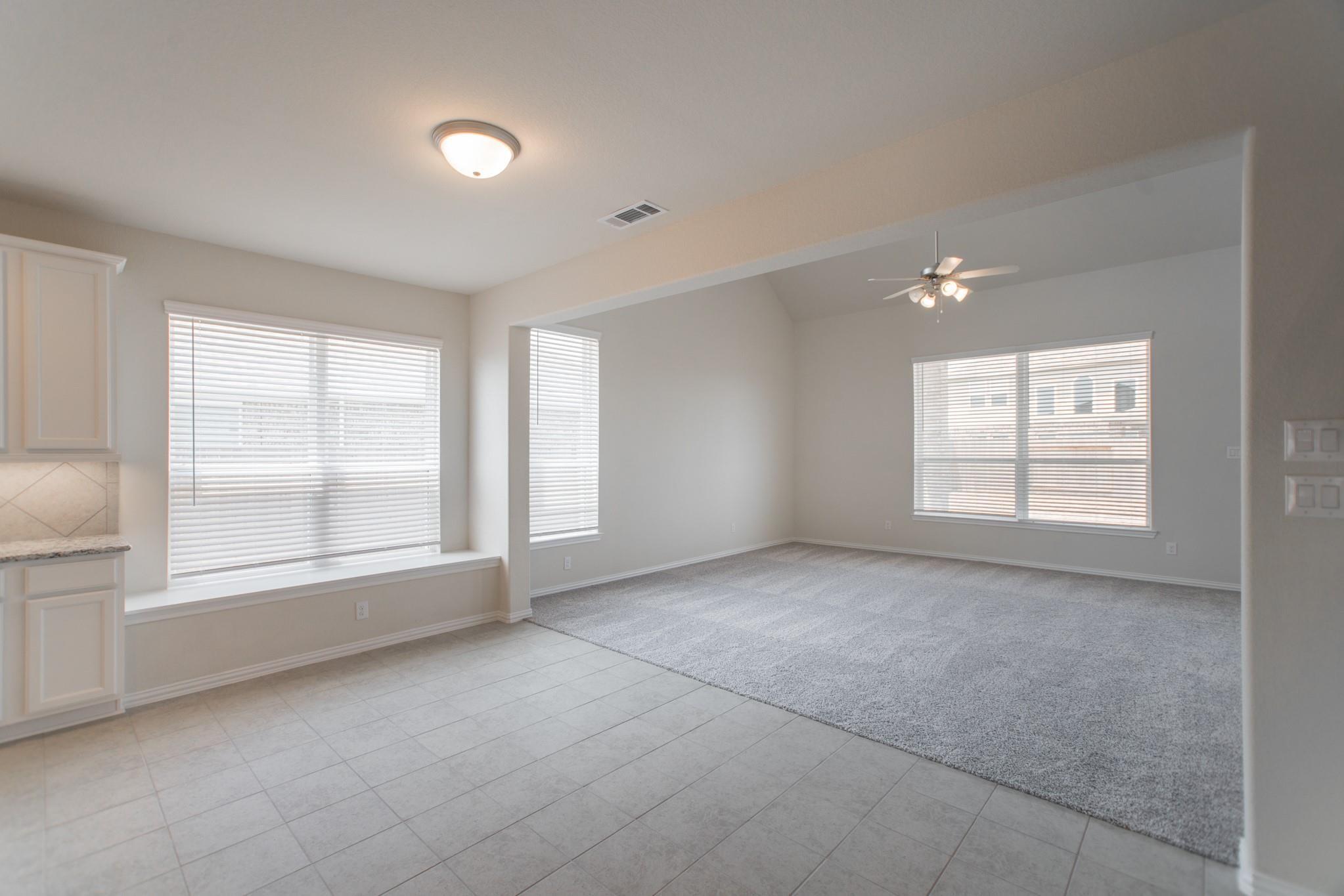 Empty living room with gray carpet, ceiling fan, and adjoining tiled area with large windows.