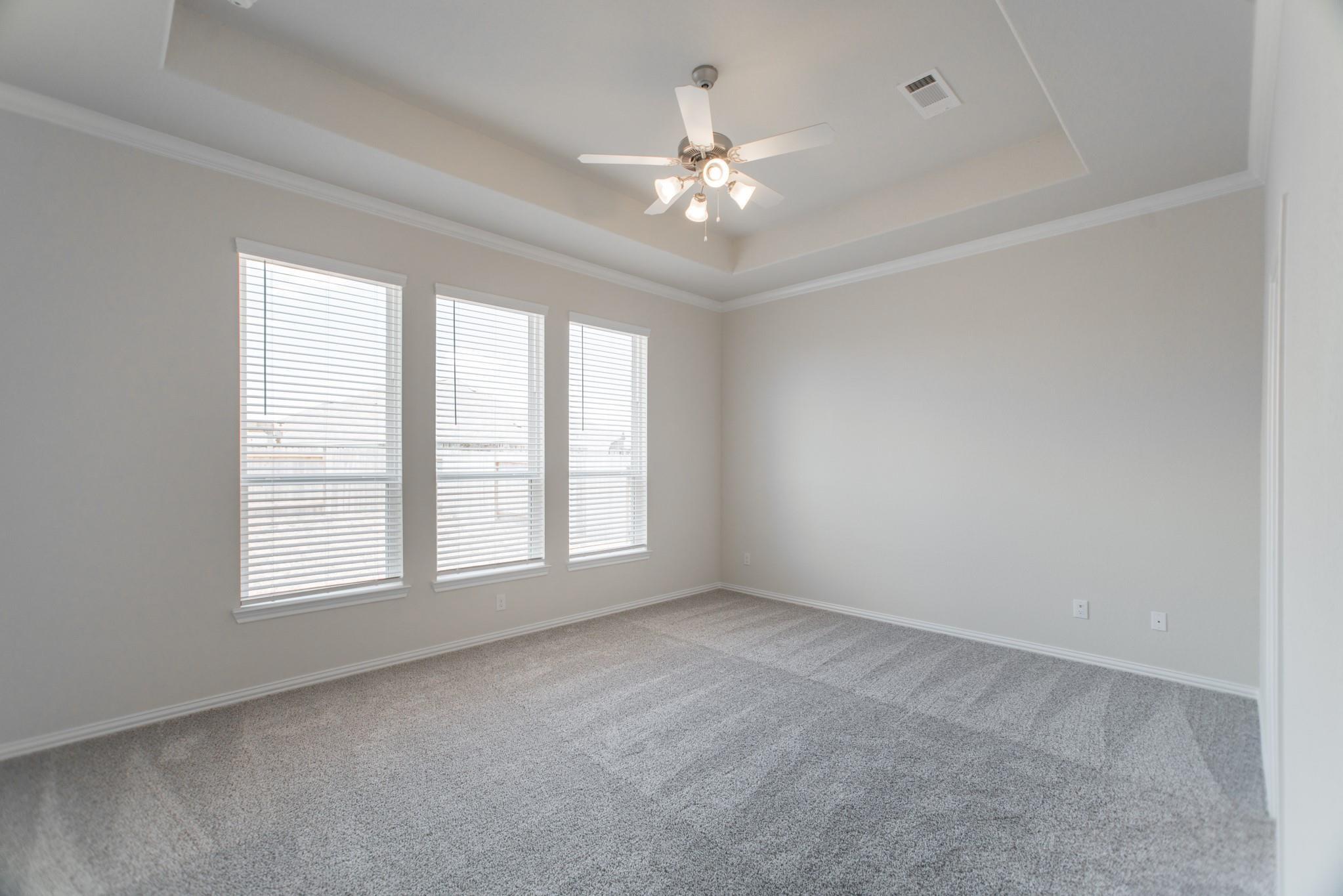 Vacant carpeted room with three blinded windows and a lit ceiling fan.
