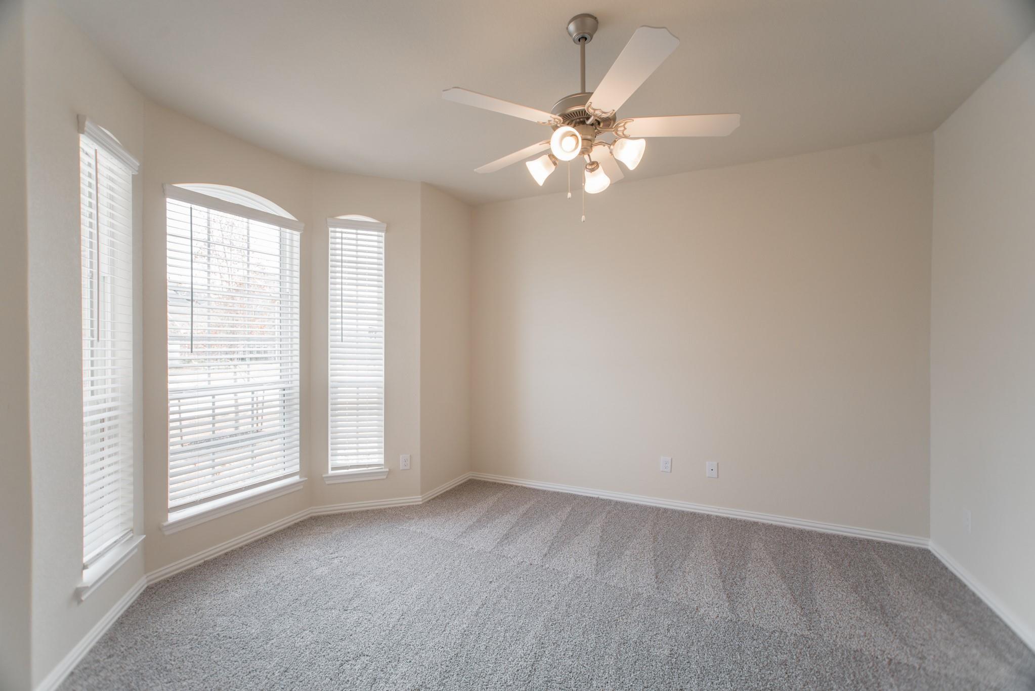 Empty carpeted bedroom with three shuttered windows and a lit ceiling fan.