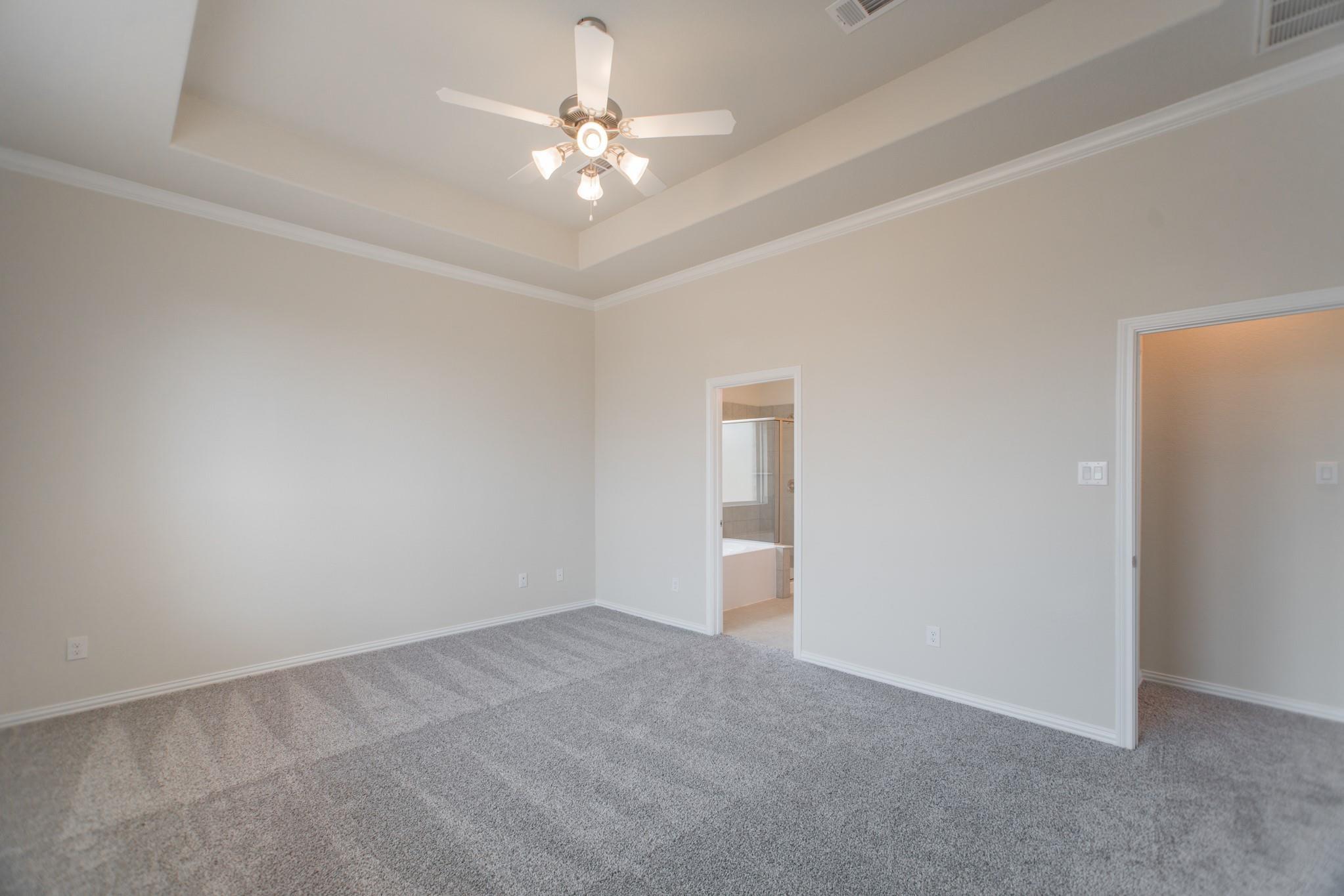 Empty bedroom with gray carpet, tray ceiling, ceiling fan, and doorway to an adjoining bathroom.