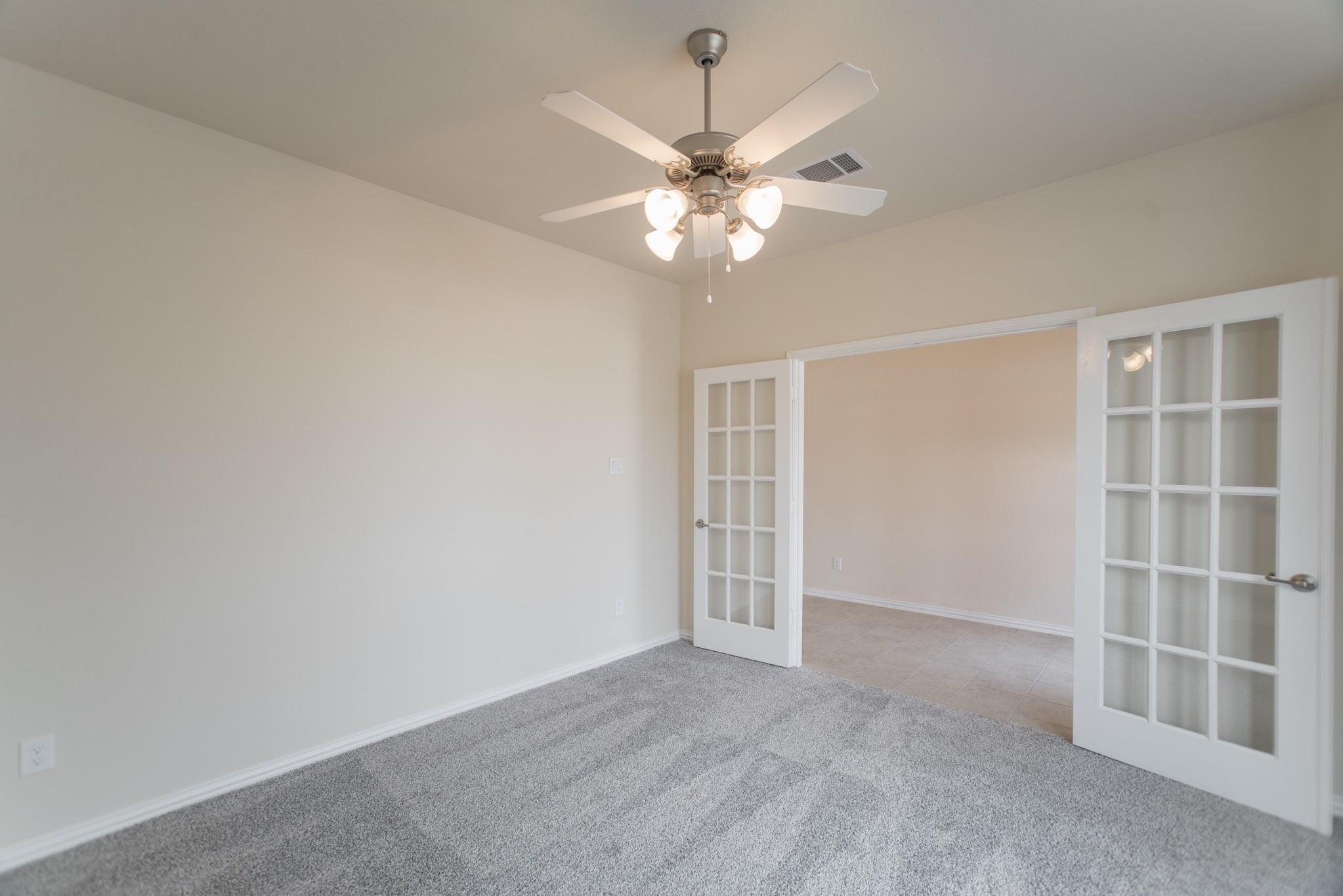 Empty carpeted room with a ceiling fan and open French doors to a tiled room.