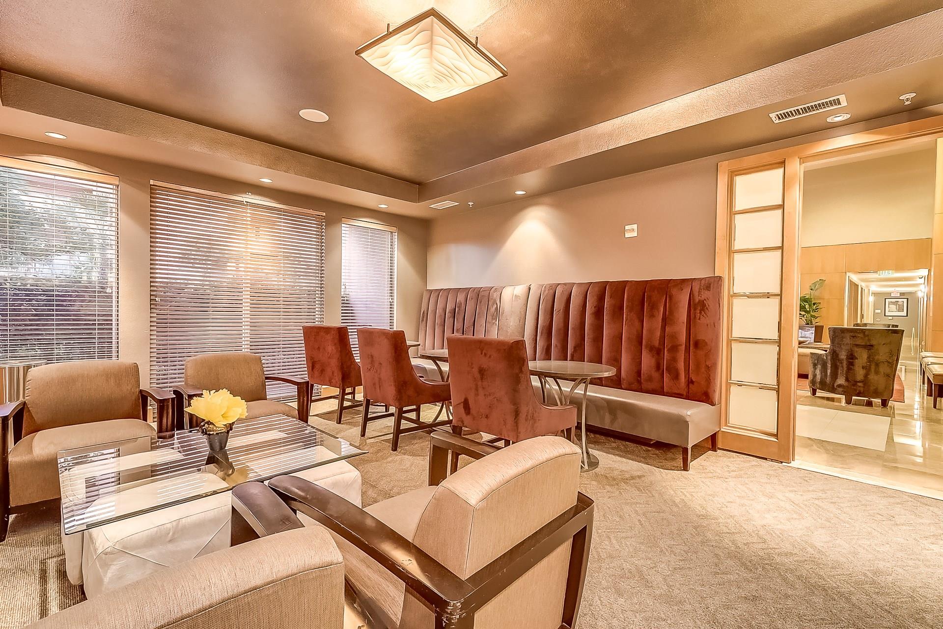 Furnished condo lounge with a burgundy banquette, armchairs, glass tables, and broad windows.