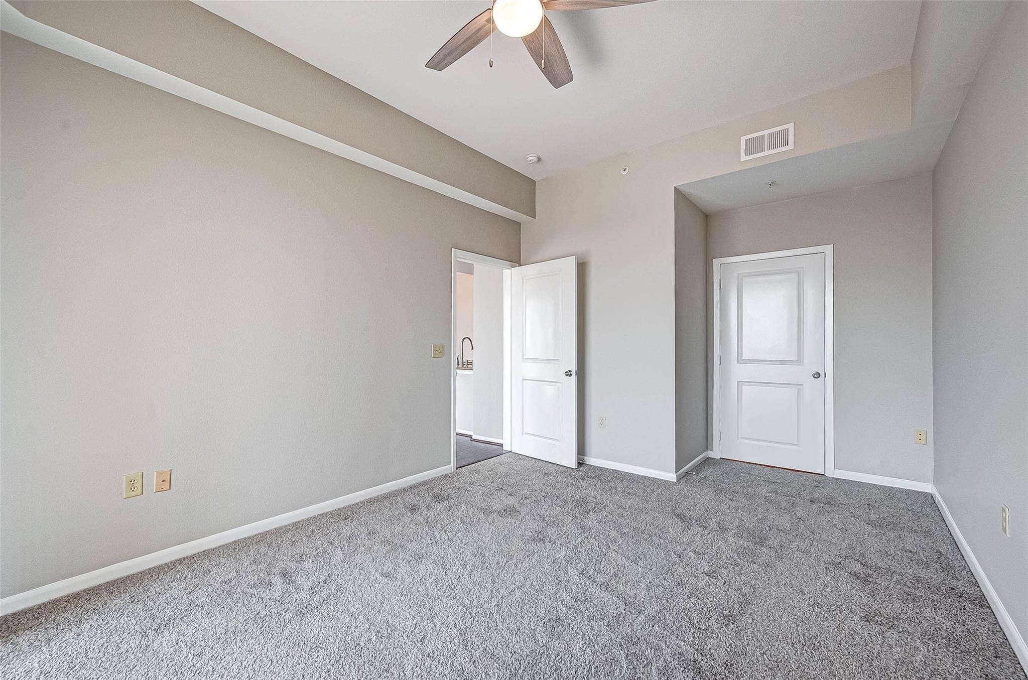 Vacant bedroom with gray carpet, ceiling fan, and an open doorway to the bathroom.