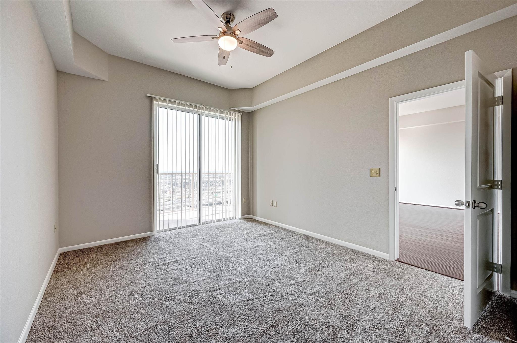 Empty carpeted bedroom with a ceiling fan, vertical blinds, and an open doorway.
