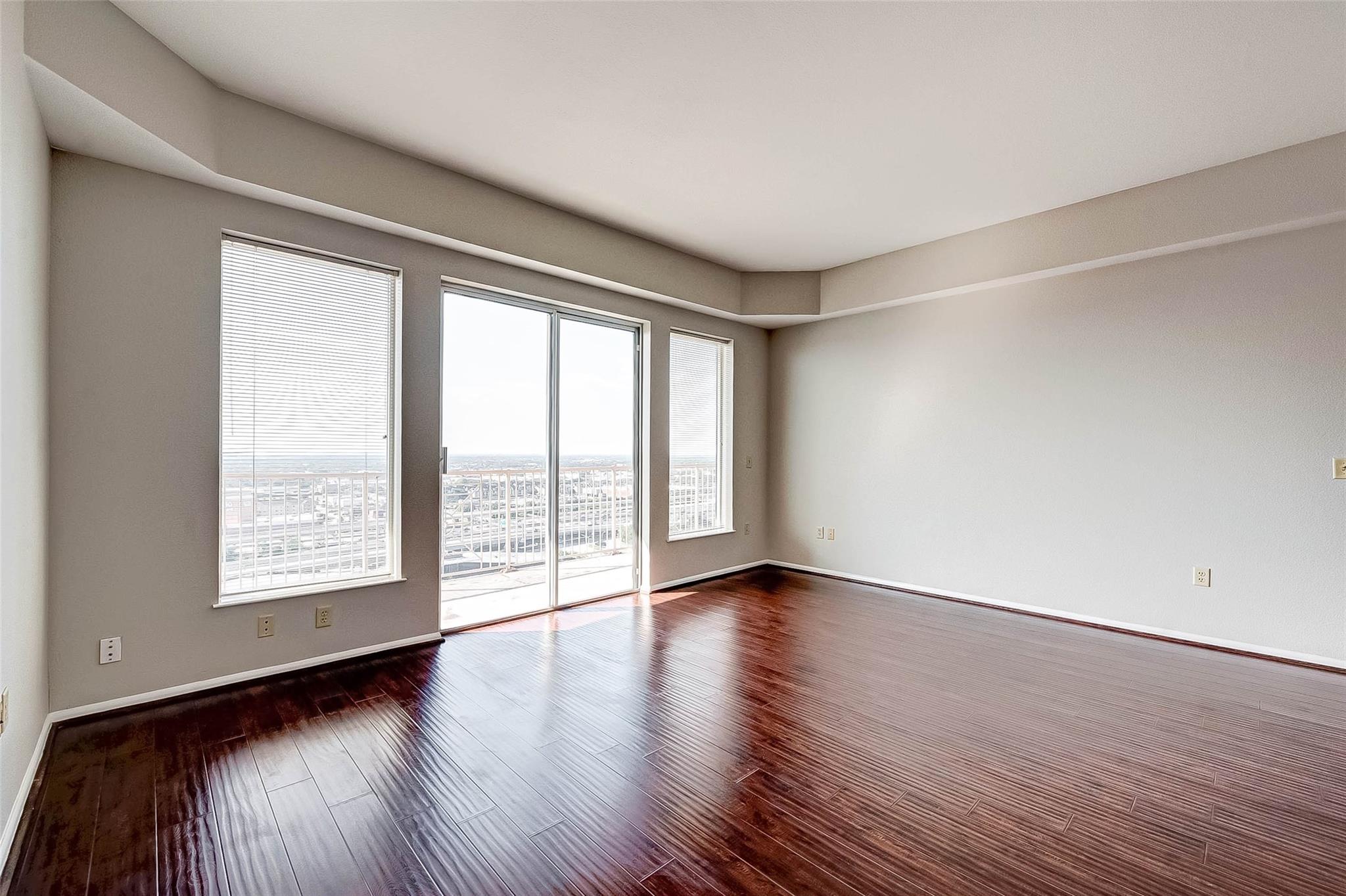 Empty condo living room with glossy dark wood floors and a balcony overlooking the city.