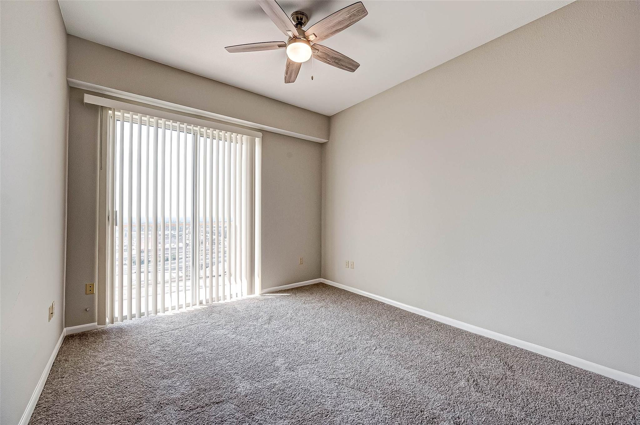 Empty carpeted bedroom with a ceiling fan and vertical blinds over a sliding glass door.
