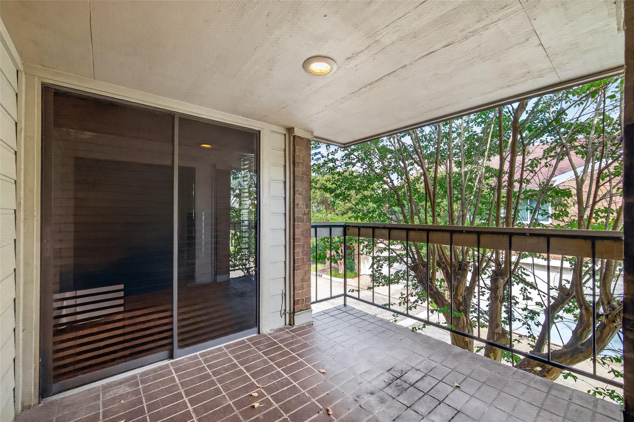 Covered tiled balcony with sliding glass door and railing overlooking leafy trees.