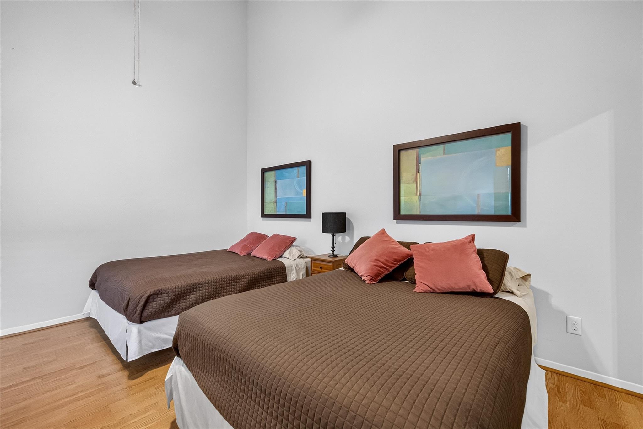 Bedroom with two brown-covered beds, coral pillows, framed art, and hardwood floors.