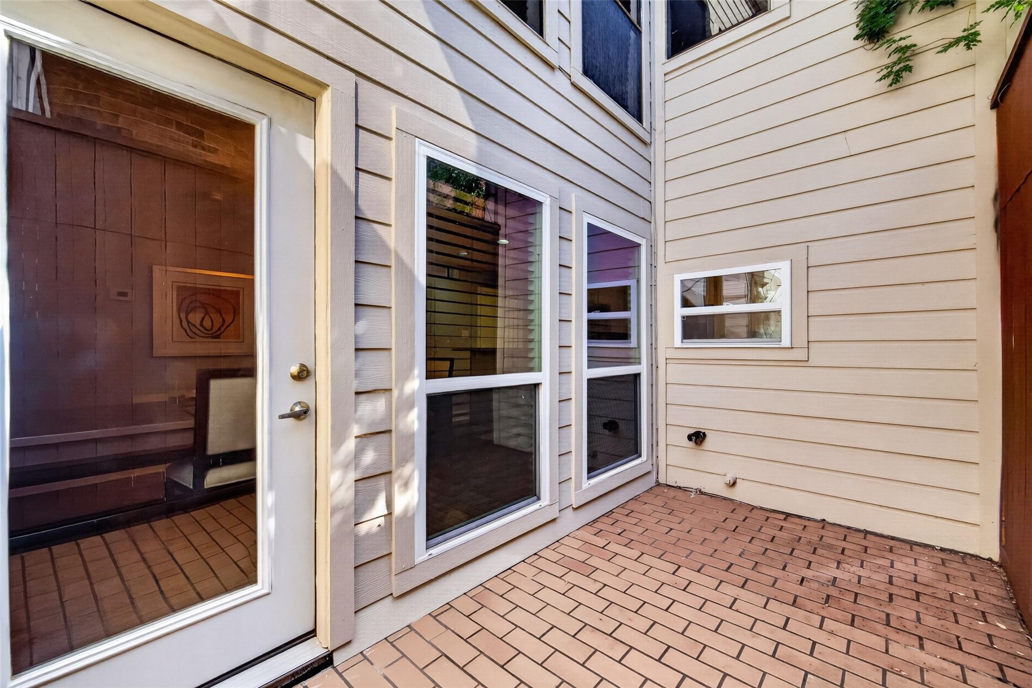 Brick-paved patio beside beige siding, with multiple windows and a glass door opening into a room.