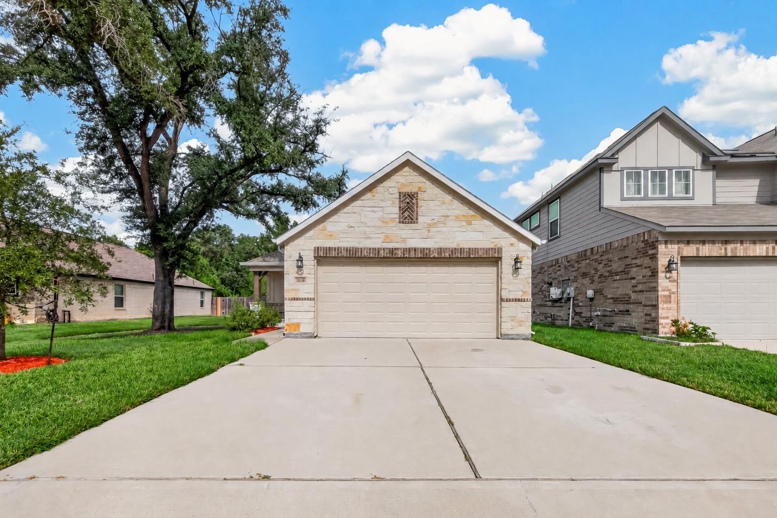 Stone-front home with an attached garage, wide driveway, lawn, and mature tree.