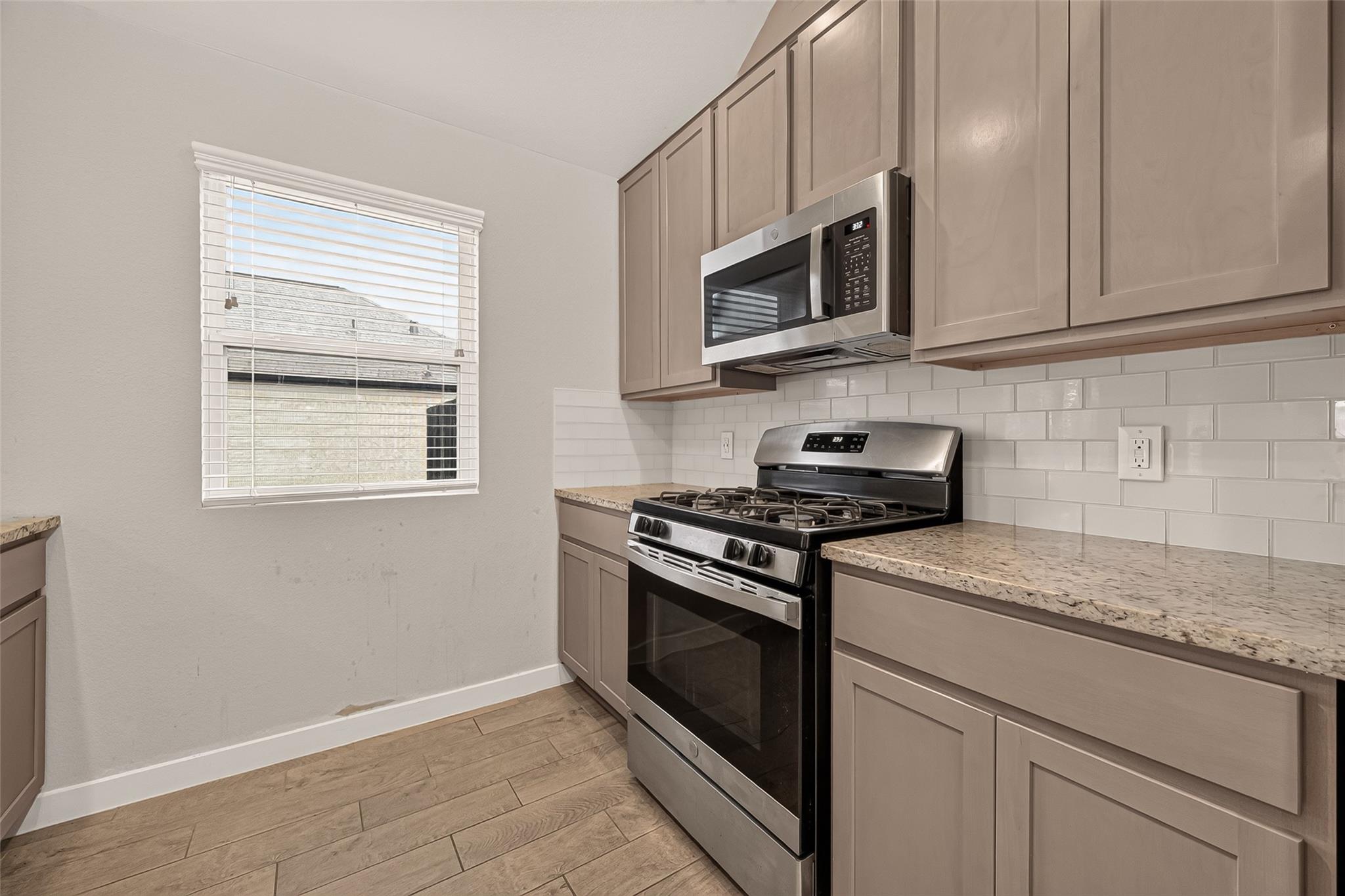 Kitchen with taupe cabinets, stainless steel gas range, subway tile backsplash, and a window with white blinds.