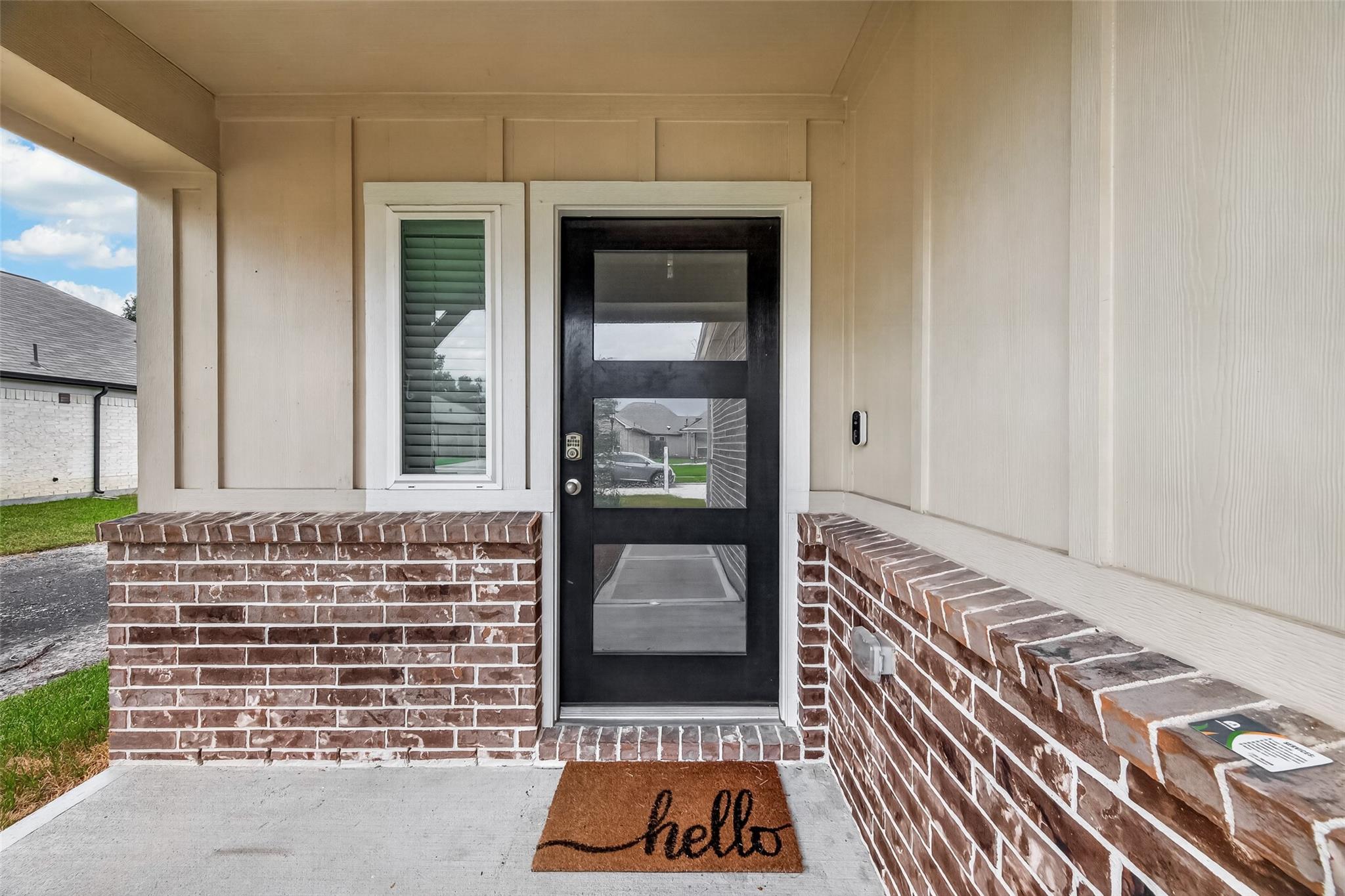 Covered house entrance with a black glass-paneled door, brick porch walls, and a “hello” doormat.