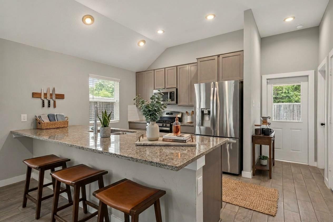 Staged kitchen with a large stone island, three bar stools, taupe cabinets, and stainless steel appliances.