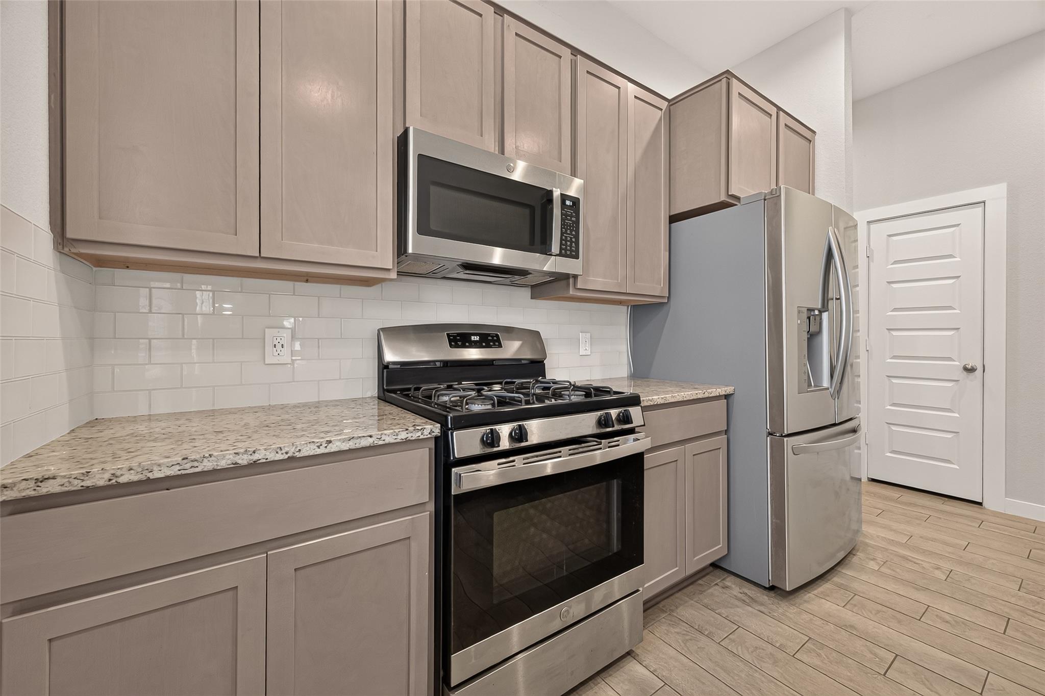 Kitchen with taupe cabinets, granite counters, stainless steel appliances, and wood-look tile flooring.