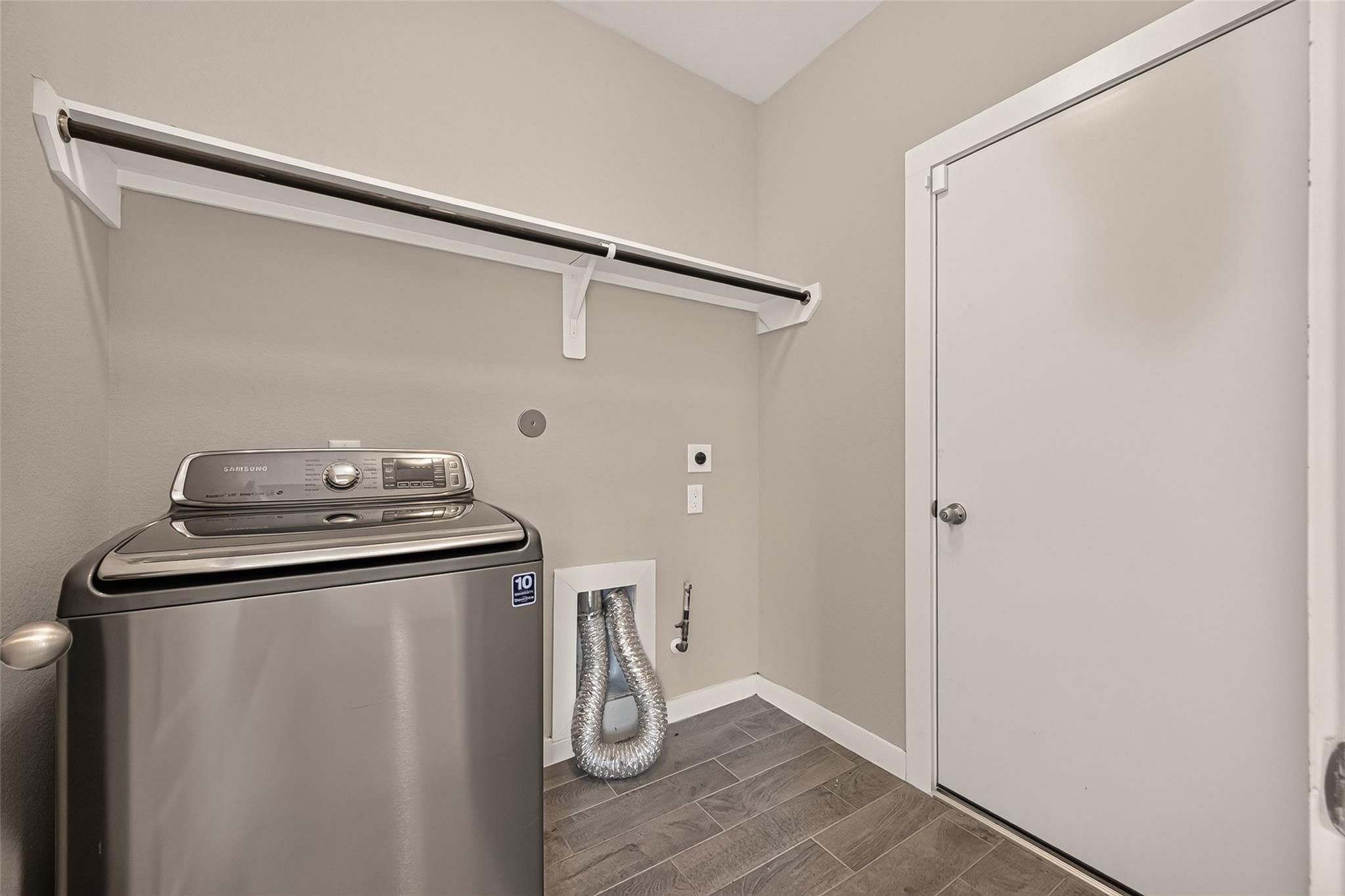 Laundry room with a Samsung washer, hanging rod, exposed vent hoses, and gray tile flooring.