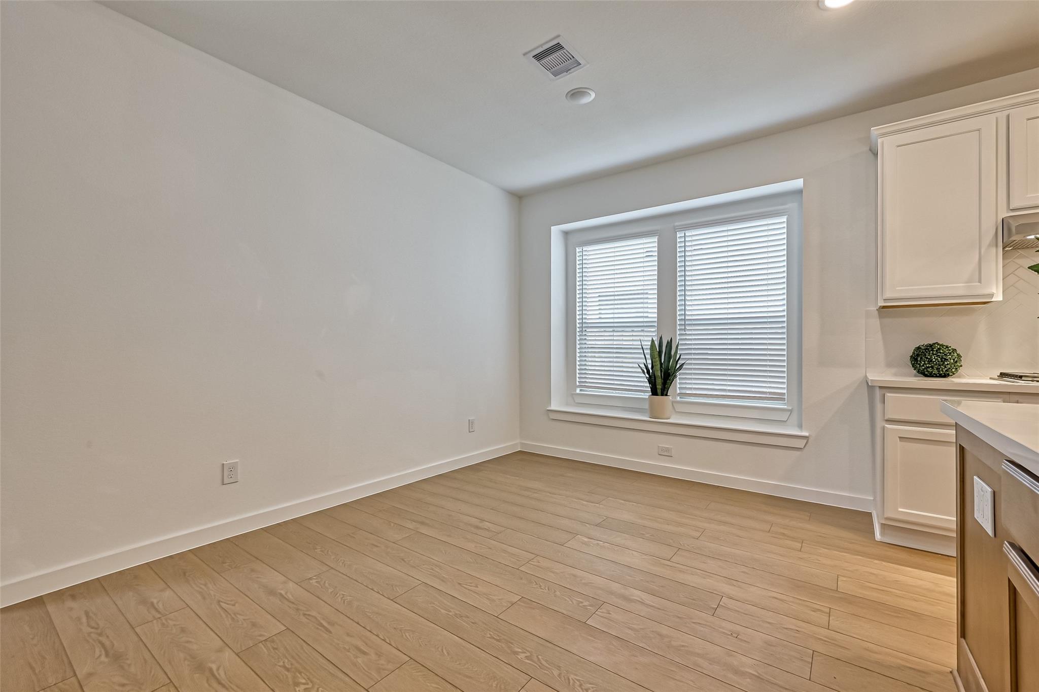 Empty dining area with light plank flooring, a wide window with blinds, and white kitchen cabinetry.