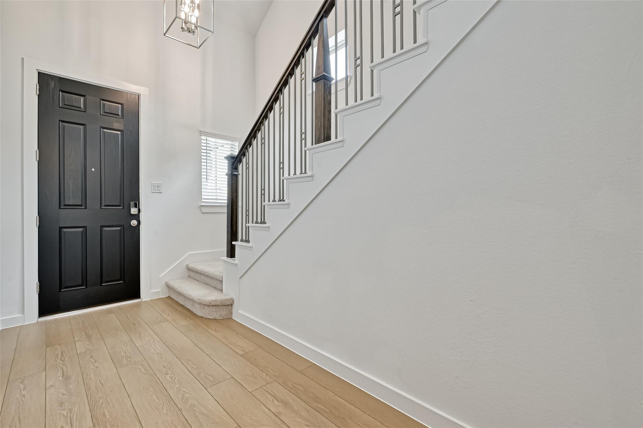 Entry foyer with a black front door, light wood flooring, and a carpeted staircase with dark railing.