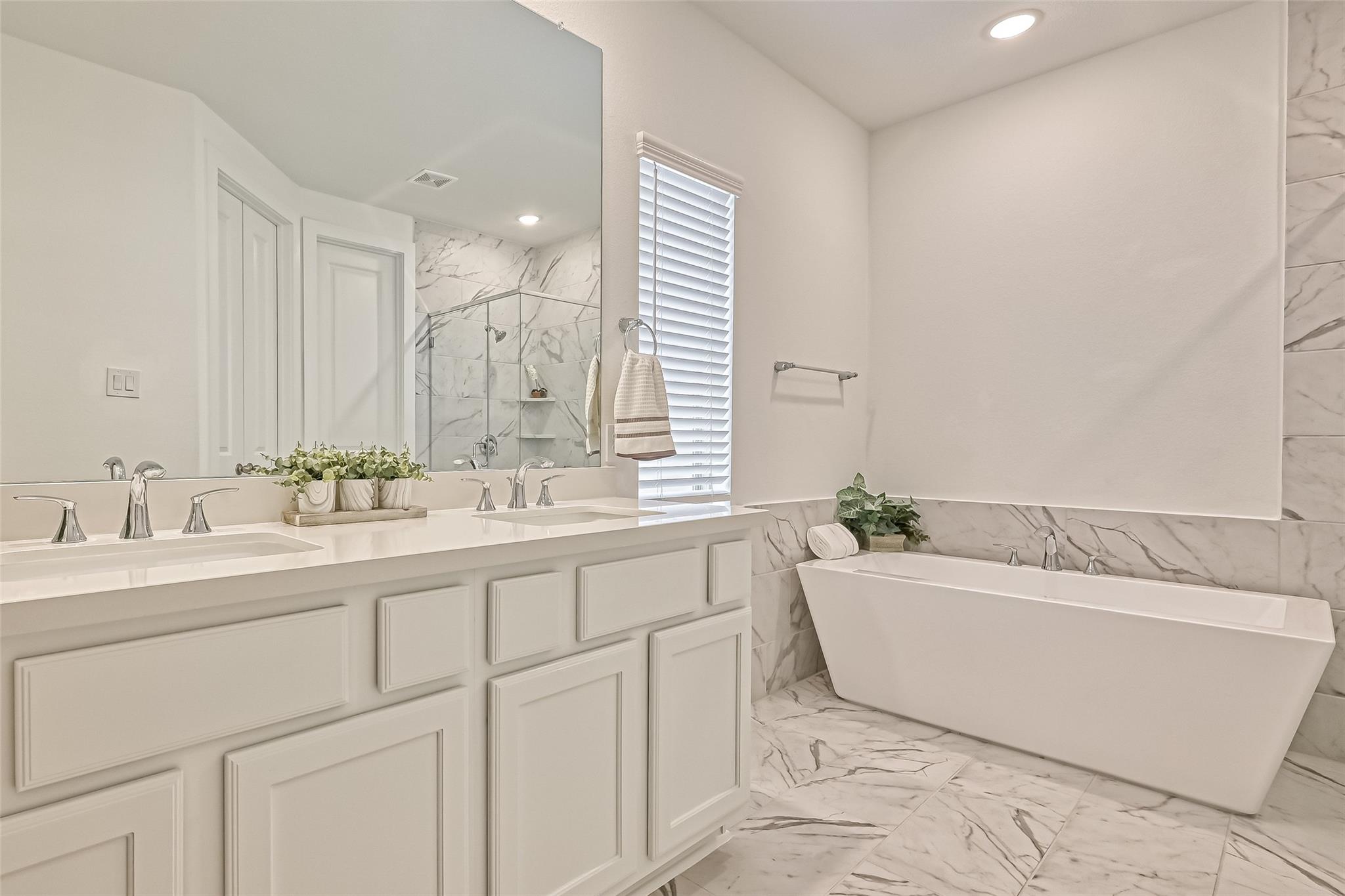 Bright bathroom with a double vanity and freestanding soaking tub beside marble-look tile.